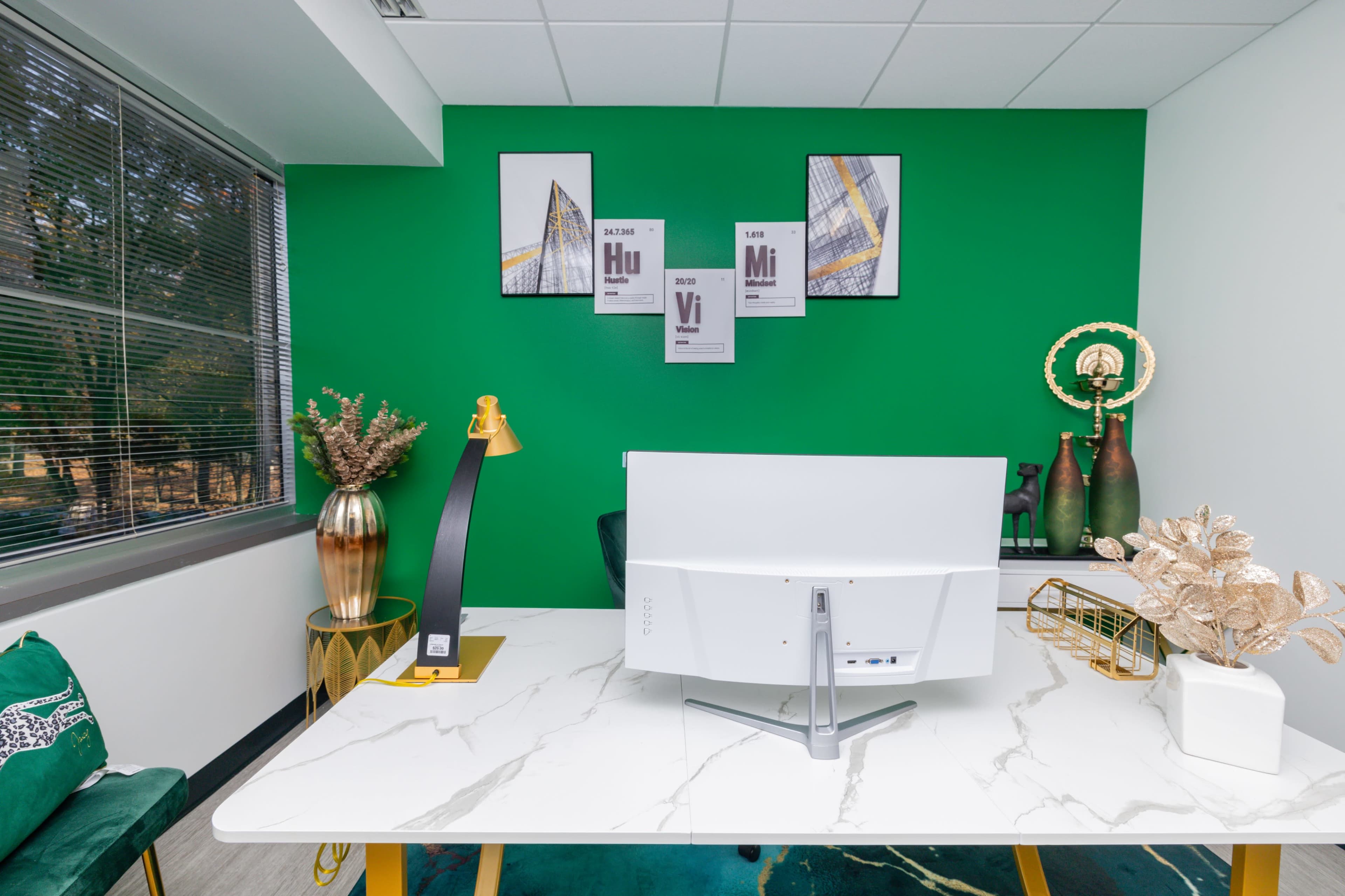 A modern office space with a green accent wall, featuring a desk with a computer monitor and decorative items, including plants and framed artwork.