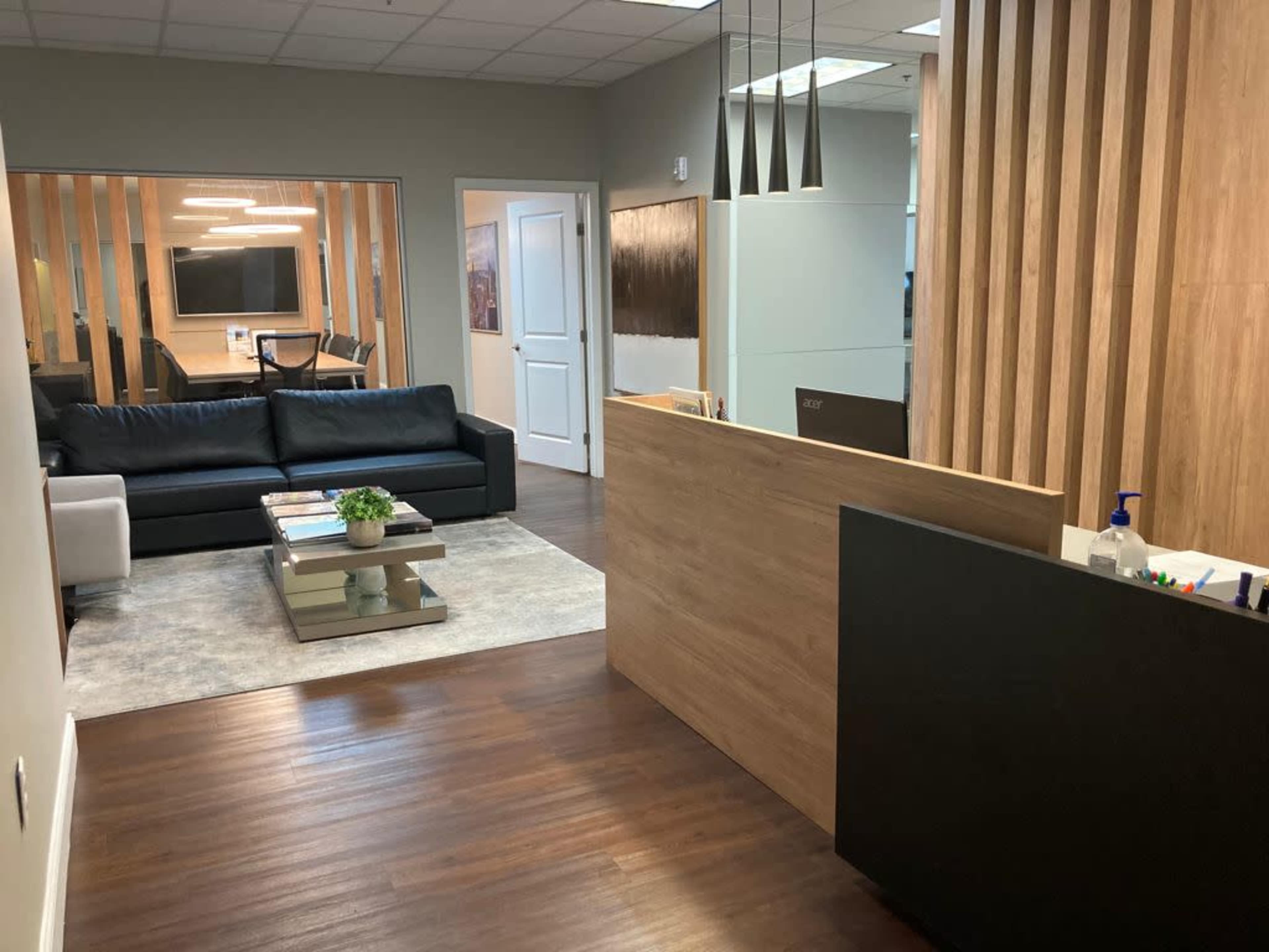 A modern office reception area with a wooden front desk, a seating area with a black couch, and a glass-walled meeting room in the background.