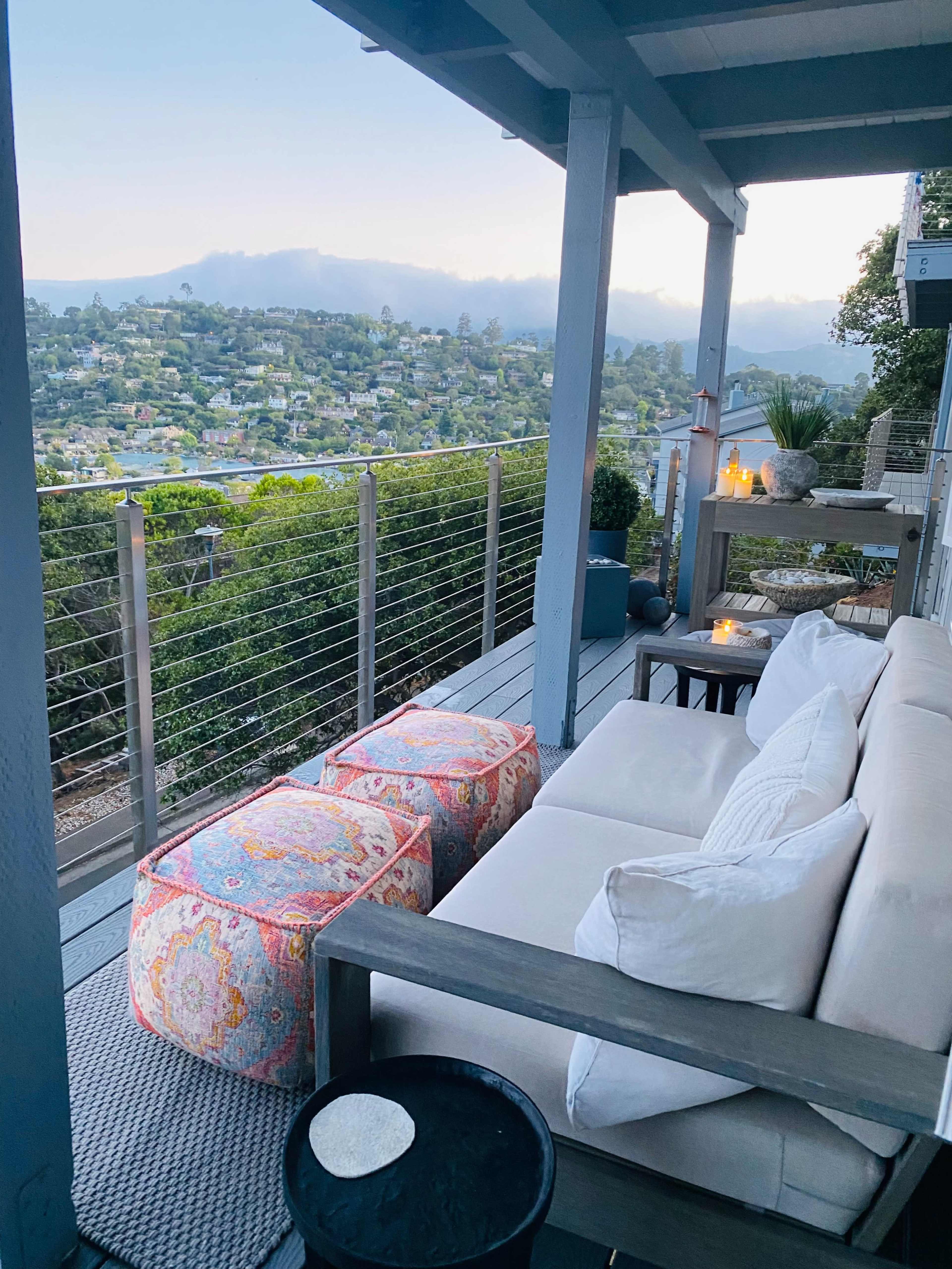 A seating area on a porch features a sofa, colorful poufs, and a view of a hillside town under a cloudy sky.