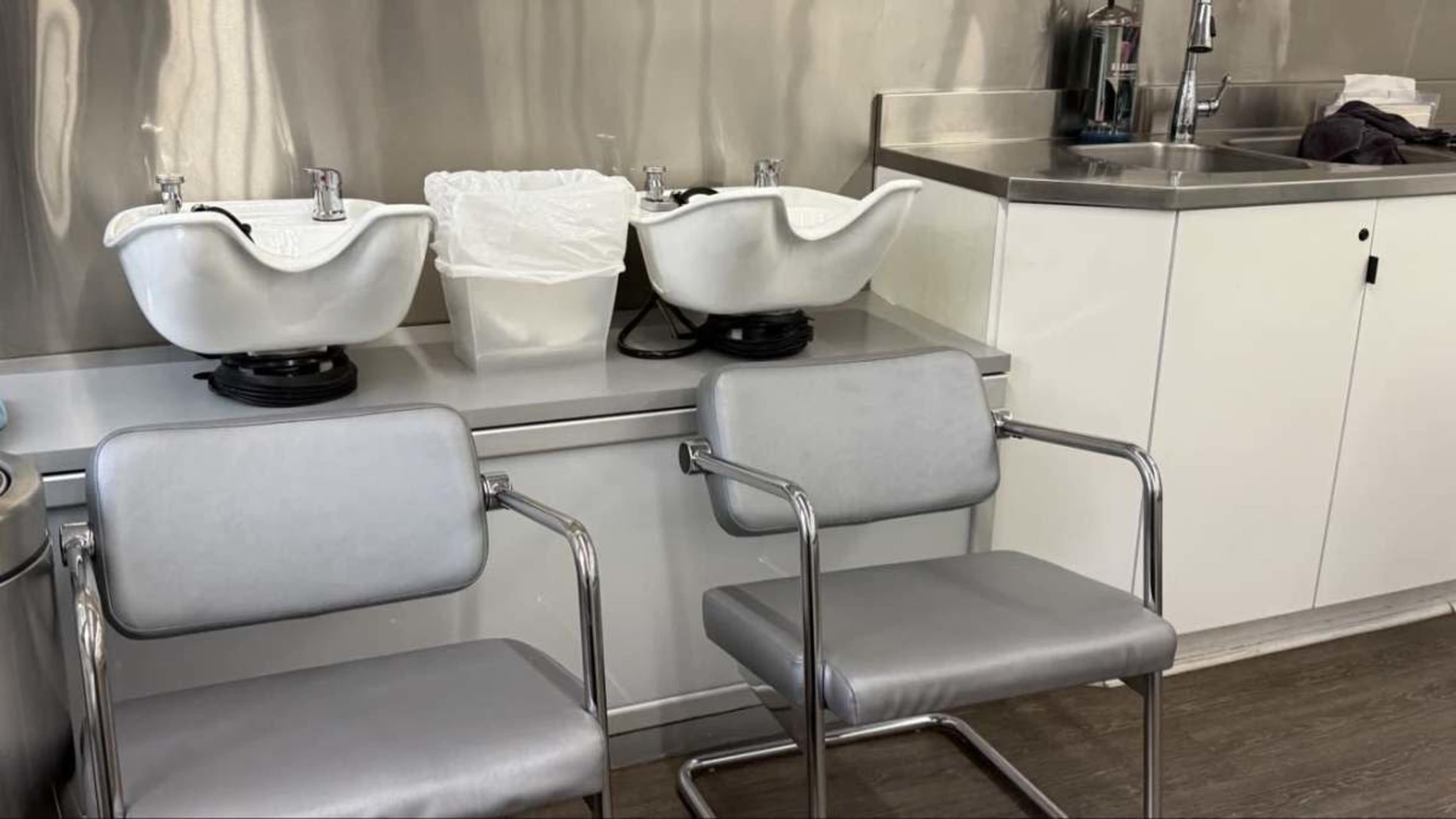 The image shows a modern salon area with two hair washing sinks, a trash bin, and two gray chairs beside a countertop with a sink and various utensils.
