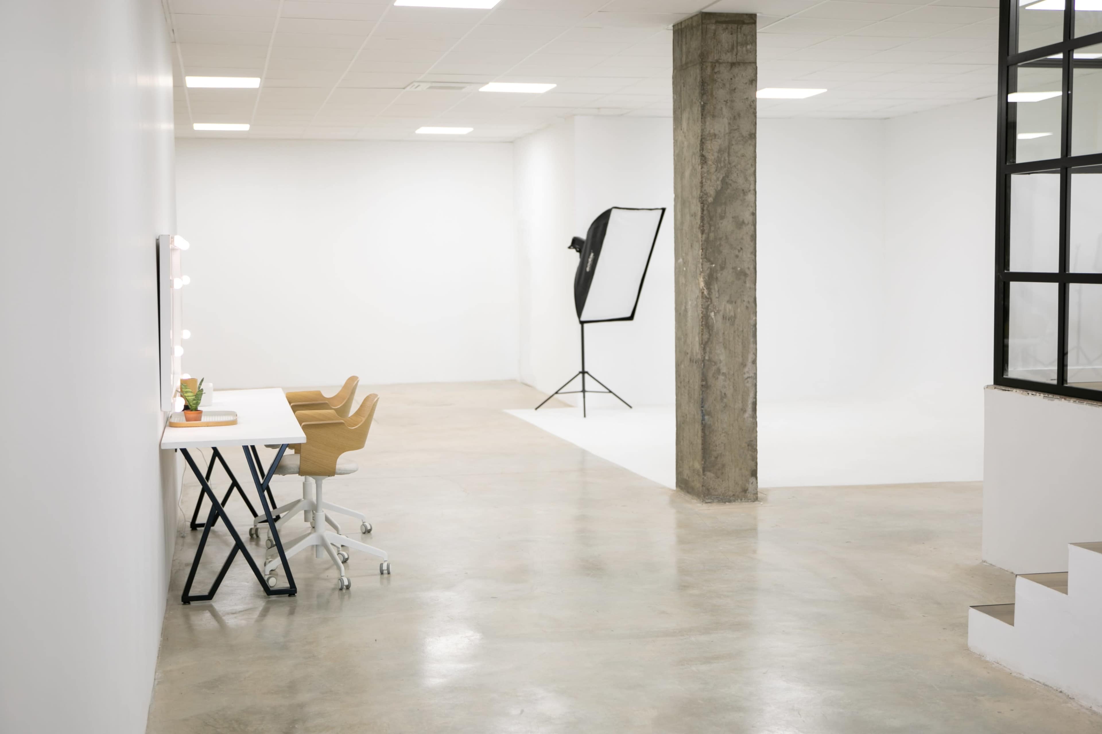 The image shows a spacious, minimalist studio featuring a white wall, polished concrete floor, two desks with chairs, and a lighting setup in the corner.