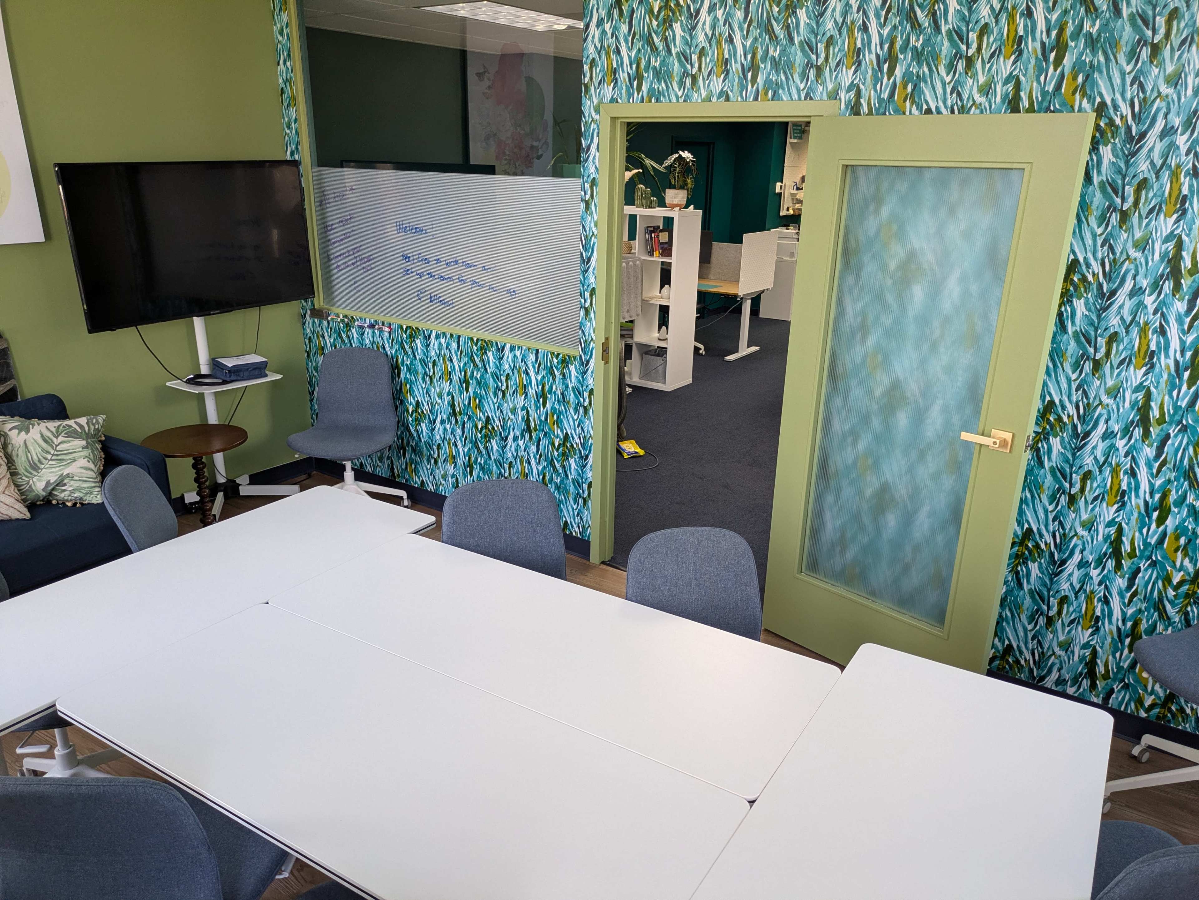 The image shows a conference room with a long white table surrounded by blue chairs, green walls, and a patterned design. A door with a glass panel leads to another room, and a television is mounted on the wall.