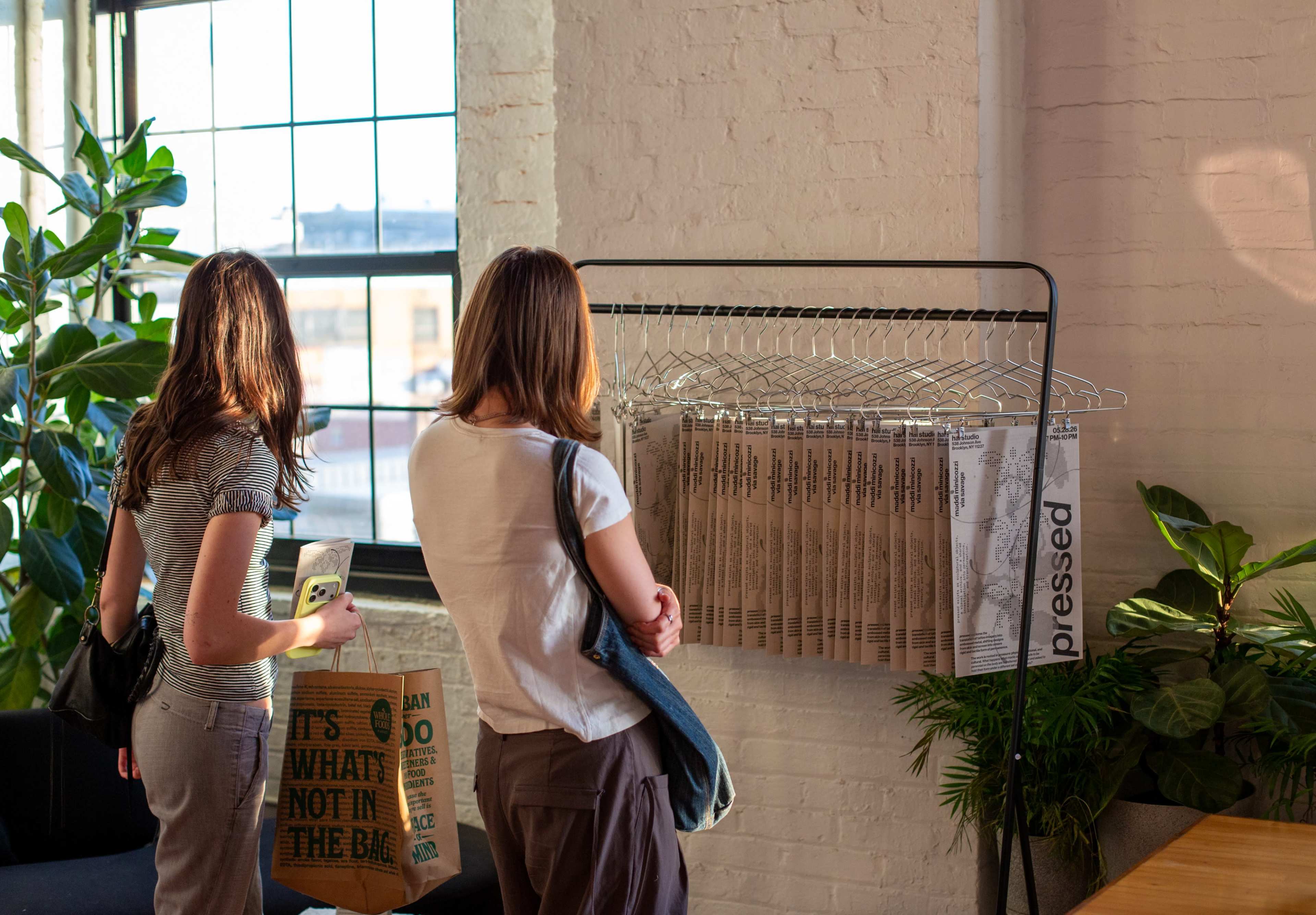 Two women stand in an urban space, examining a rack of printed materials displayed nearby large windows and greenery.