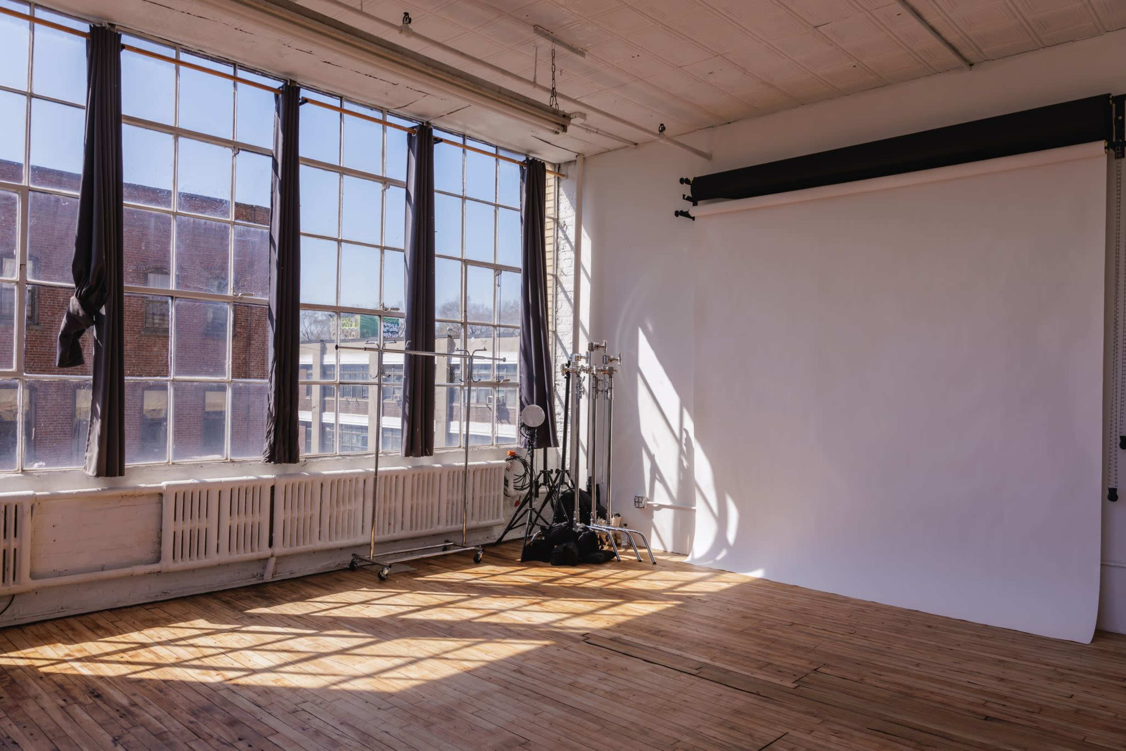 A bright photography studio features large windows, wooden flooring, and a white backdrop with lighting equipment positioned nearby.