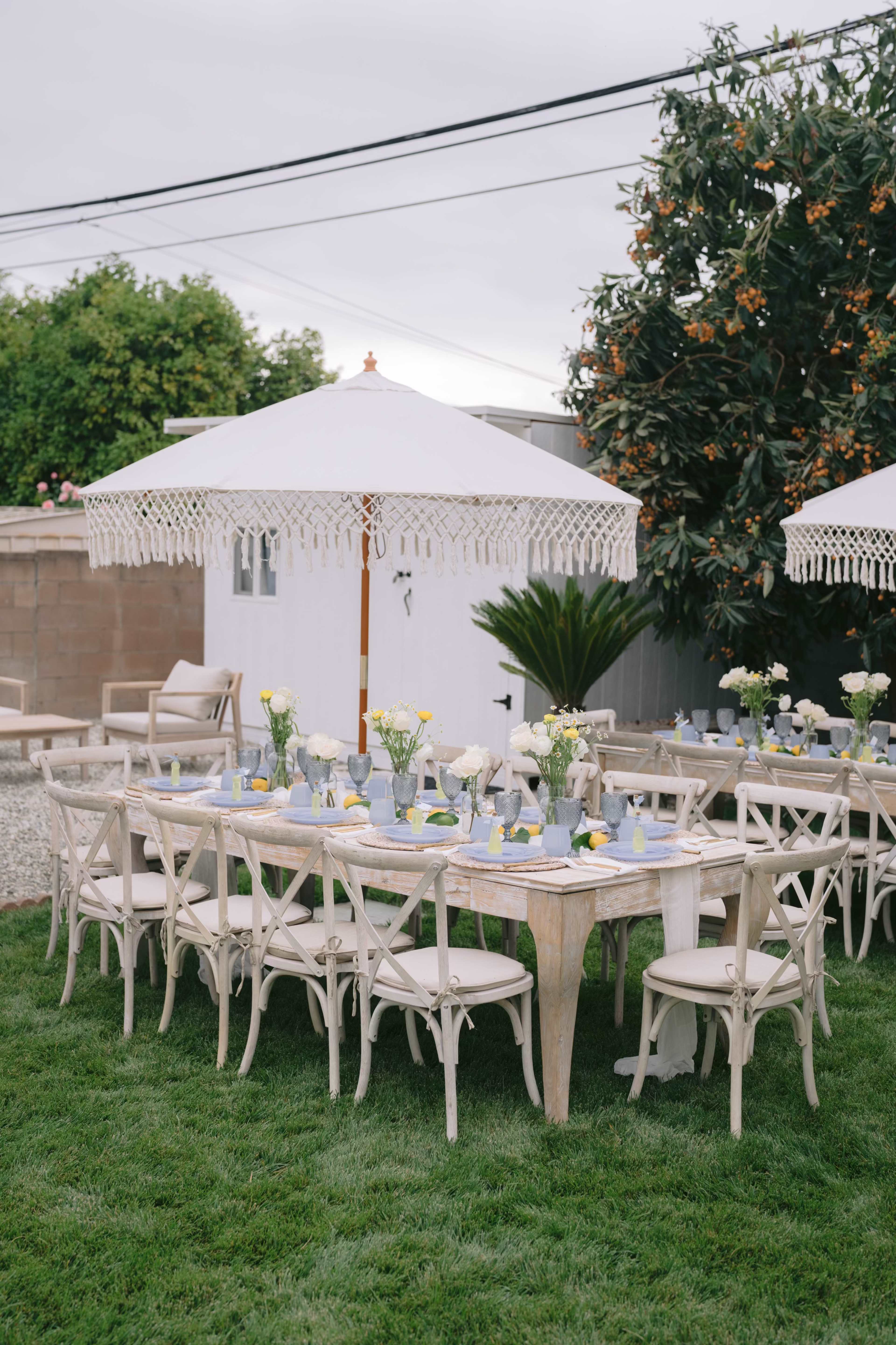 A decorated outdoor dining area features long tables with floral centerpieces, surrounded by white chairs and large umbrellas, set on green grass.