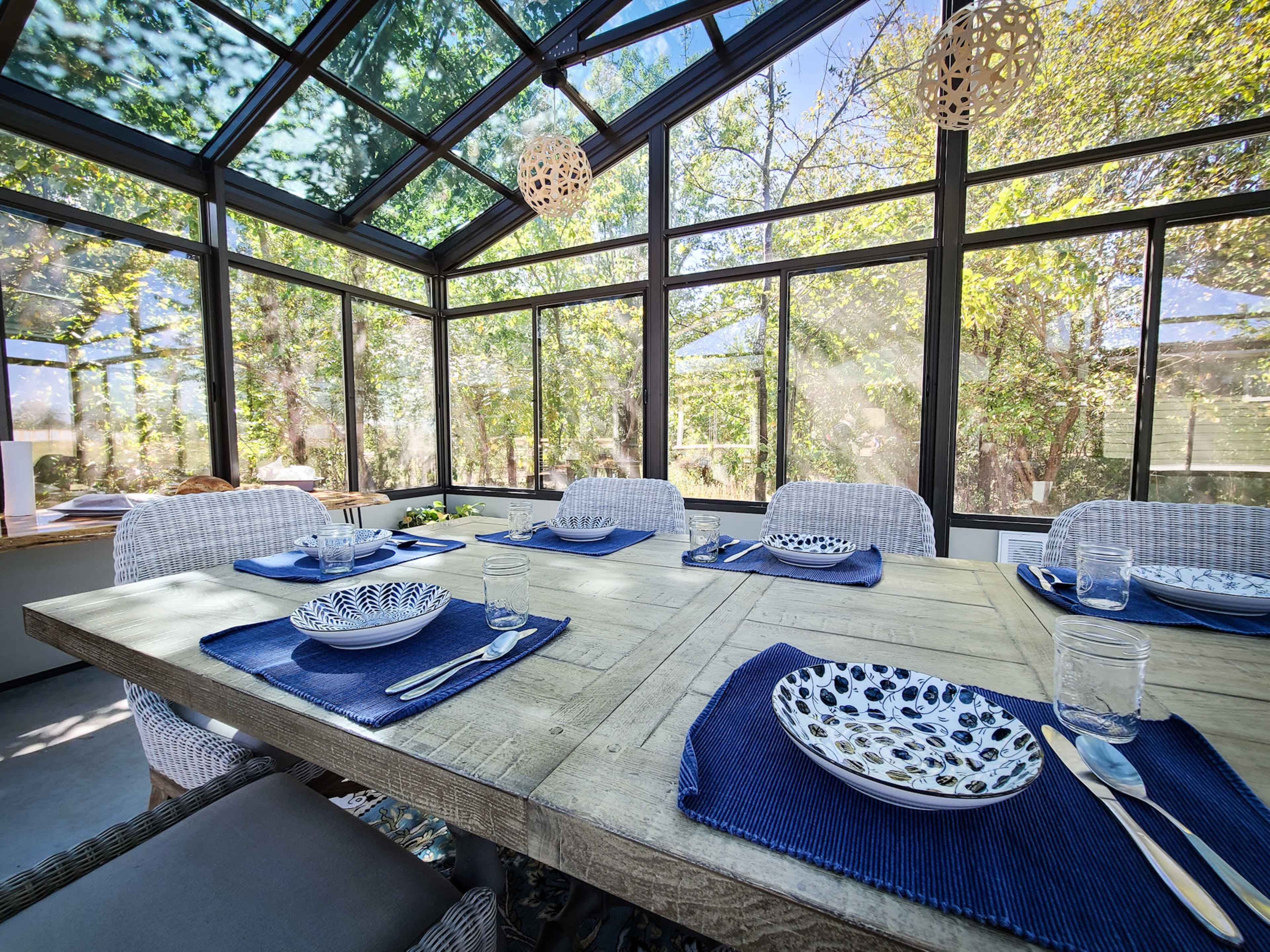 A spacious dining area features a wooden table set with decorative plates and glassware, surrounded by large windows that provide a view of the greenery outside.