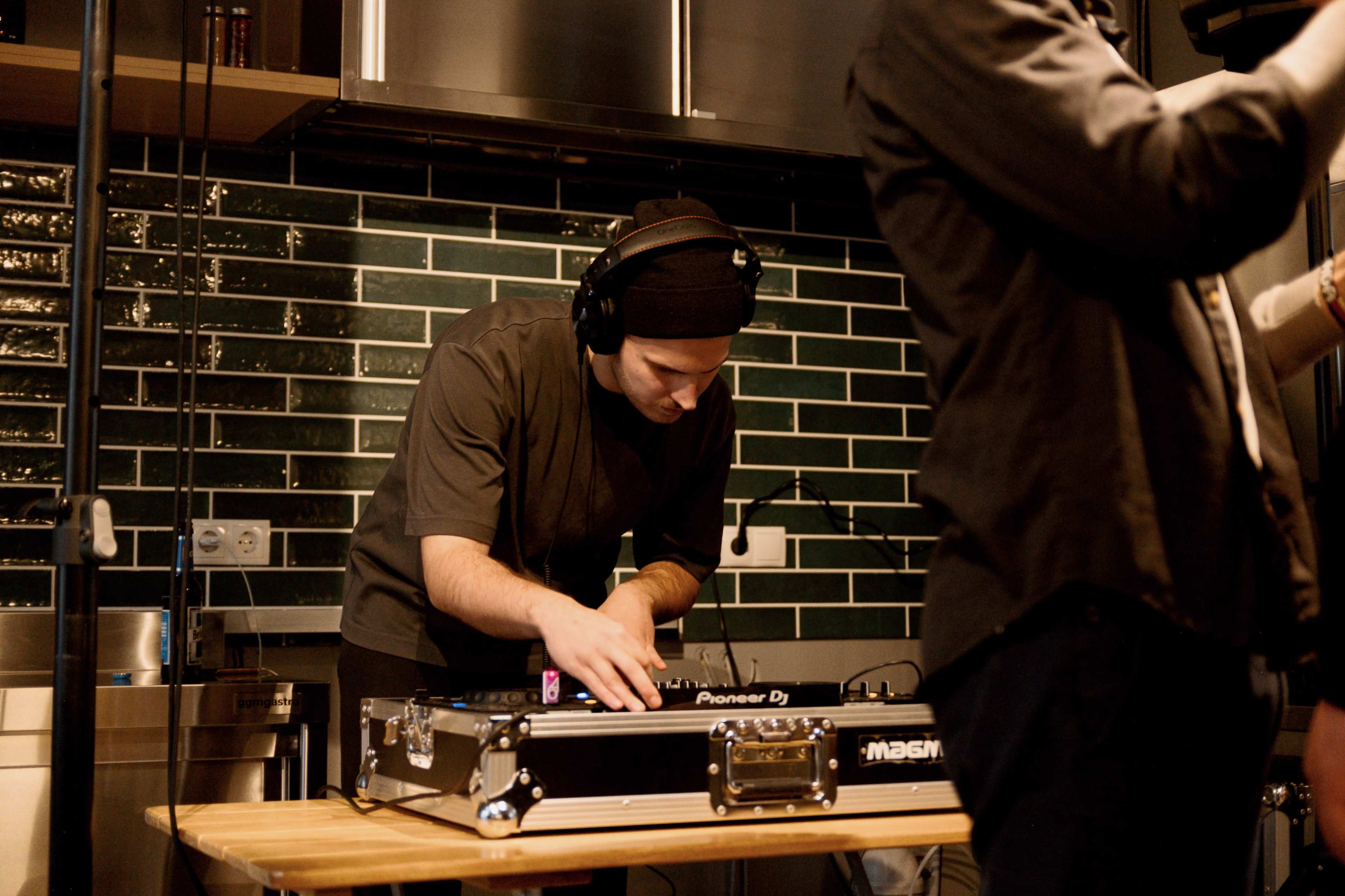 A DJ is adjusting equipment on a table in a kitchen setting with green tiled walls.