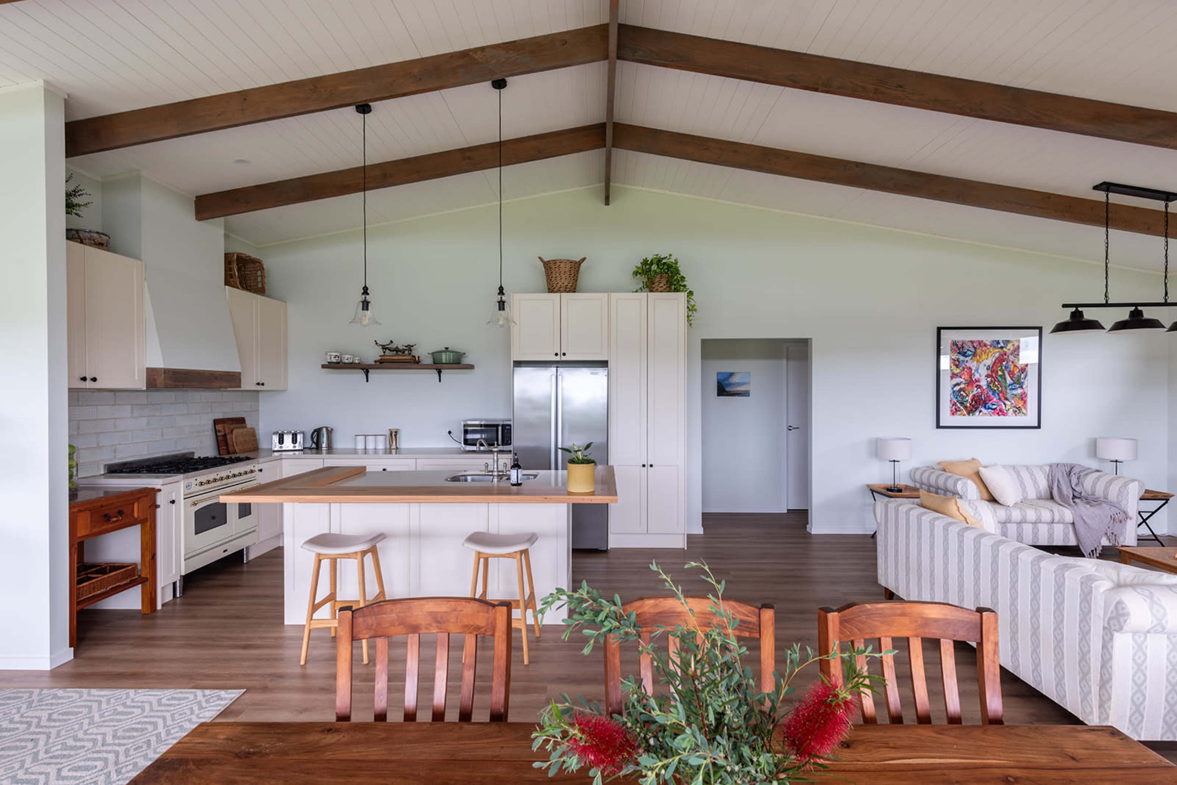 The image shows a modern kitchen and dining area with wooden beams, a central island, and a comfortable living space featuring a sofa and a dining table.