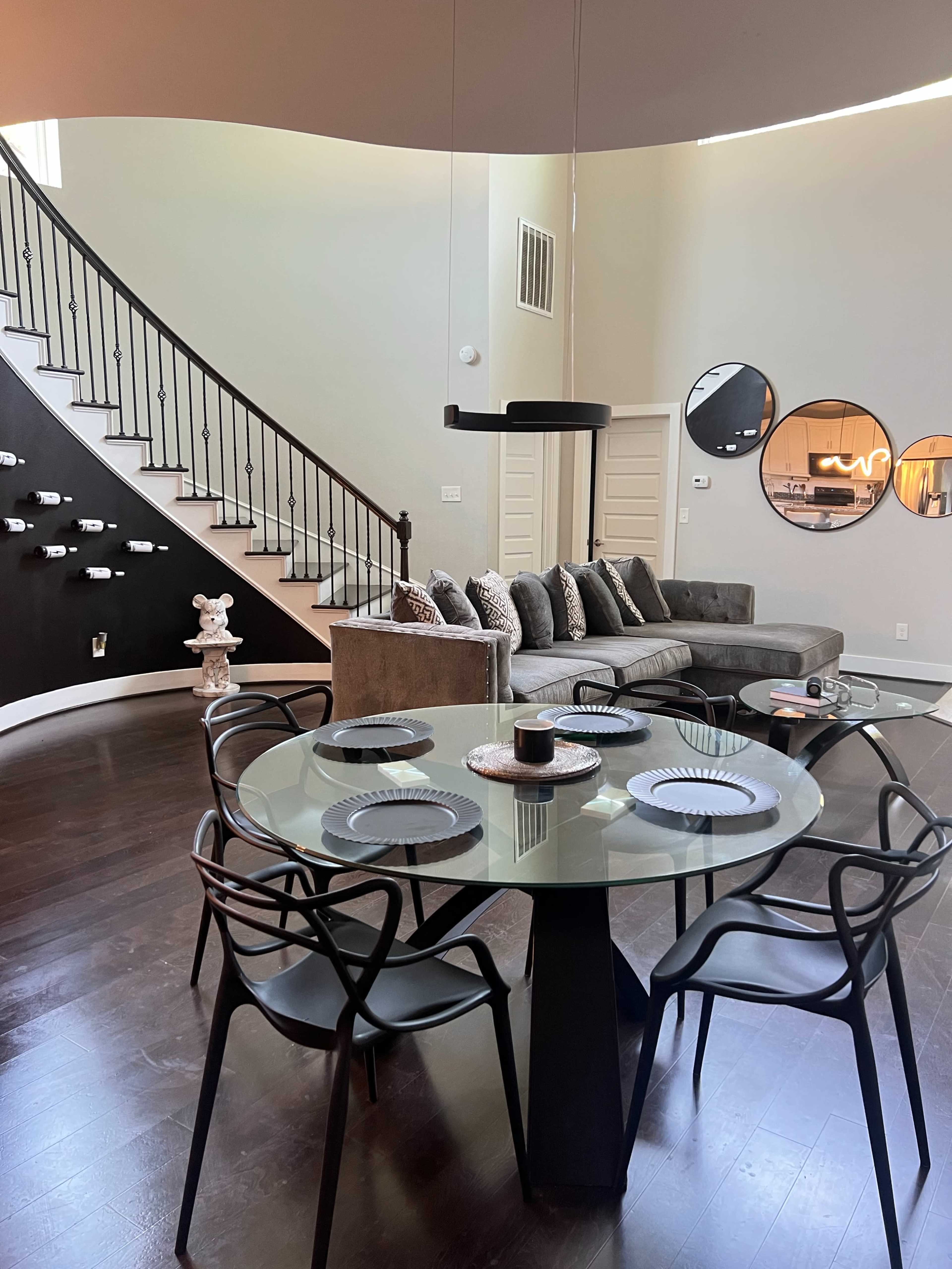 A modern living space features a dining area with a glass table and black chairs adjacent to a curved staircase leading to a sitting area with a sectional couch and a decorative wall.