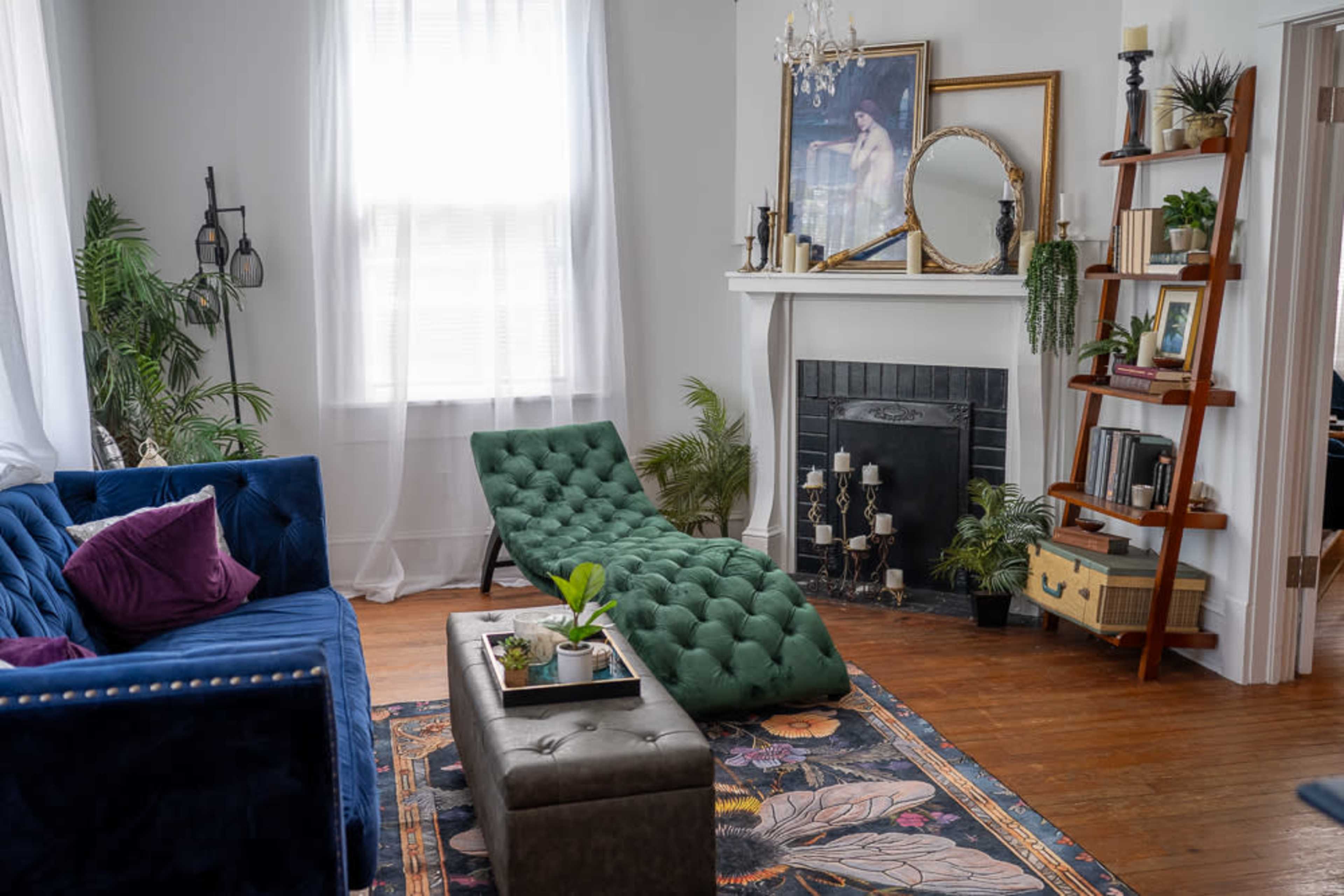 The living room features a blue velvet sofa, a green chaise lounge, a decorative fireplace, and a wooden bookshelf adorned with plants and books.