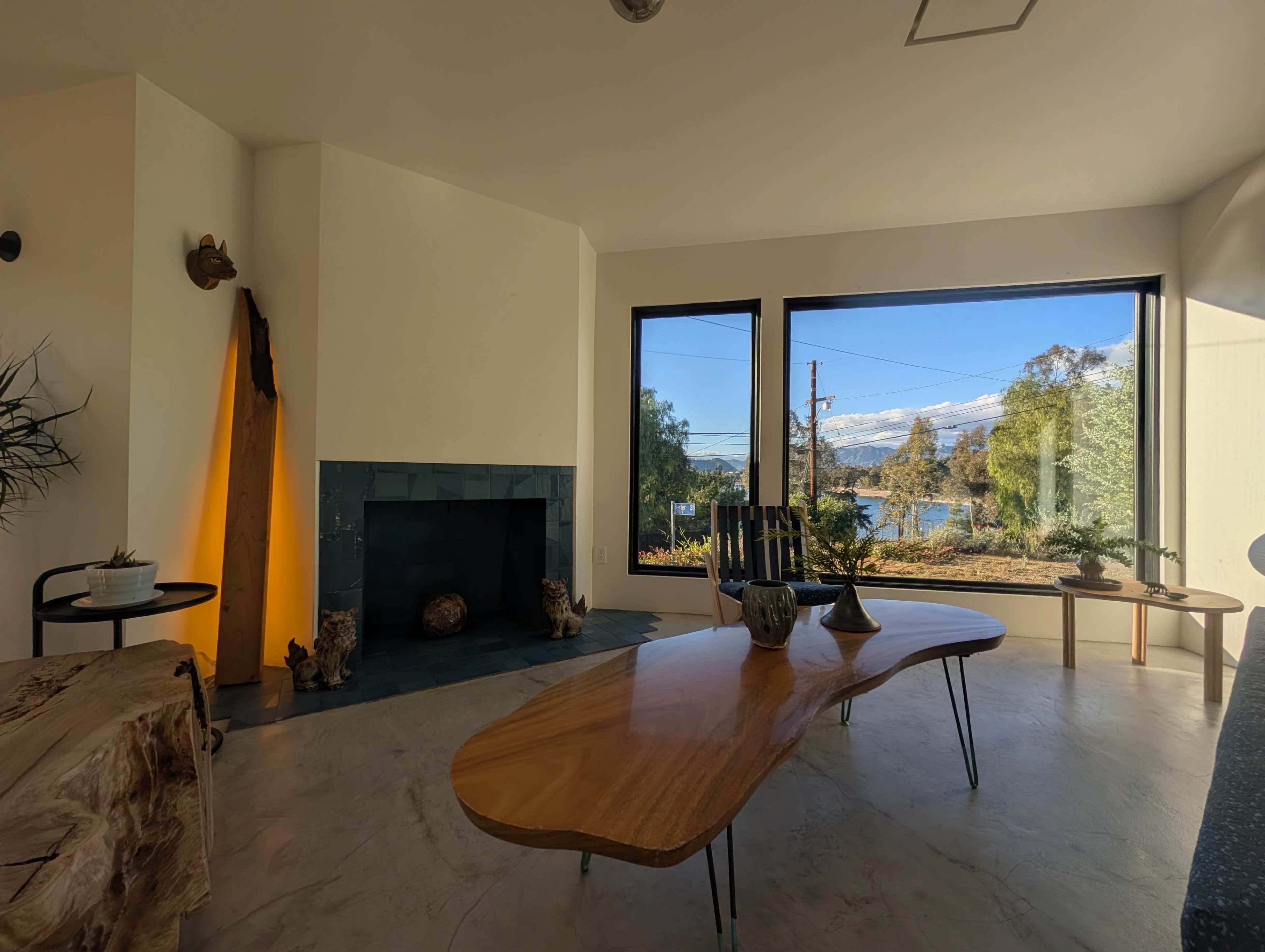 The image shows a modern interior with large windows overlooking a scenic view, featuring a wooden table, a fireplace, and minimalistic furnishings.