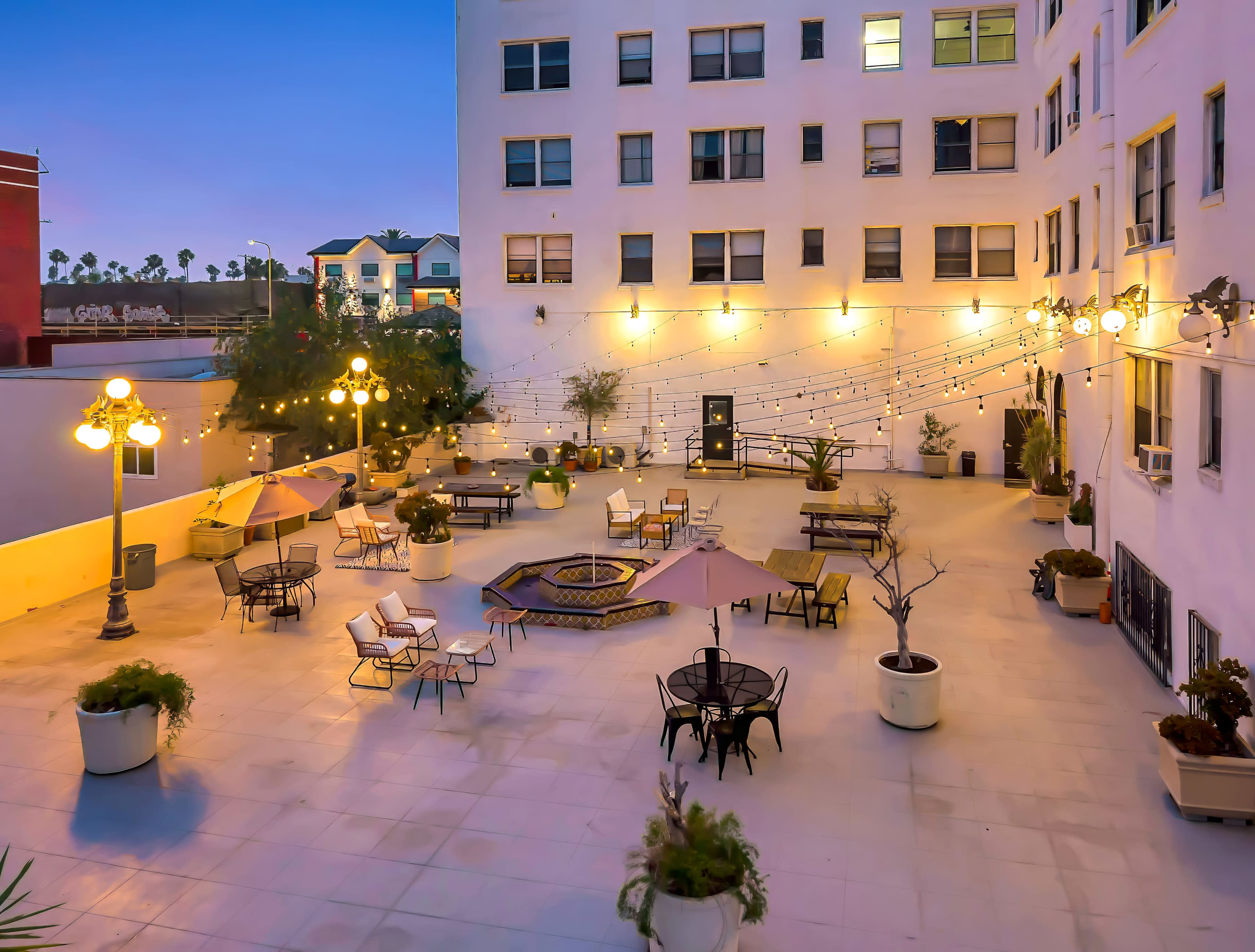 The image shows a courtyard with outdoor seating arrangements, string lights, and a central fire pit, surrounded by buildings.
