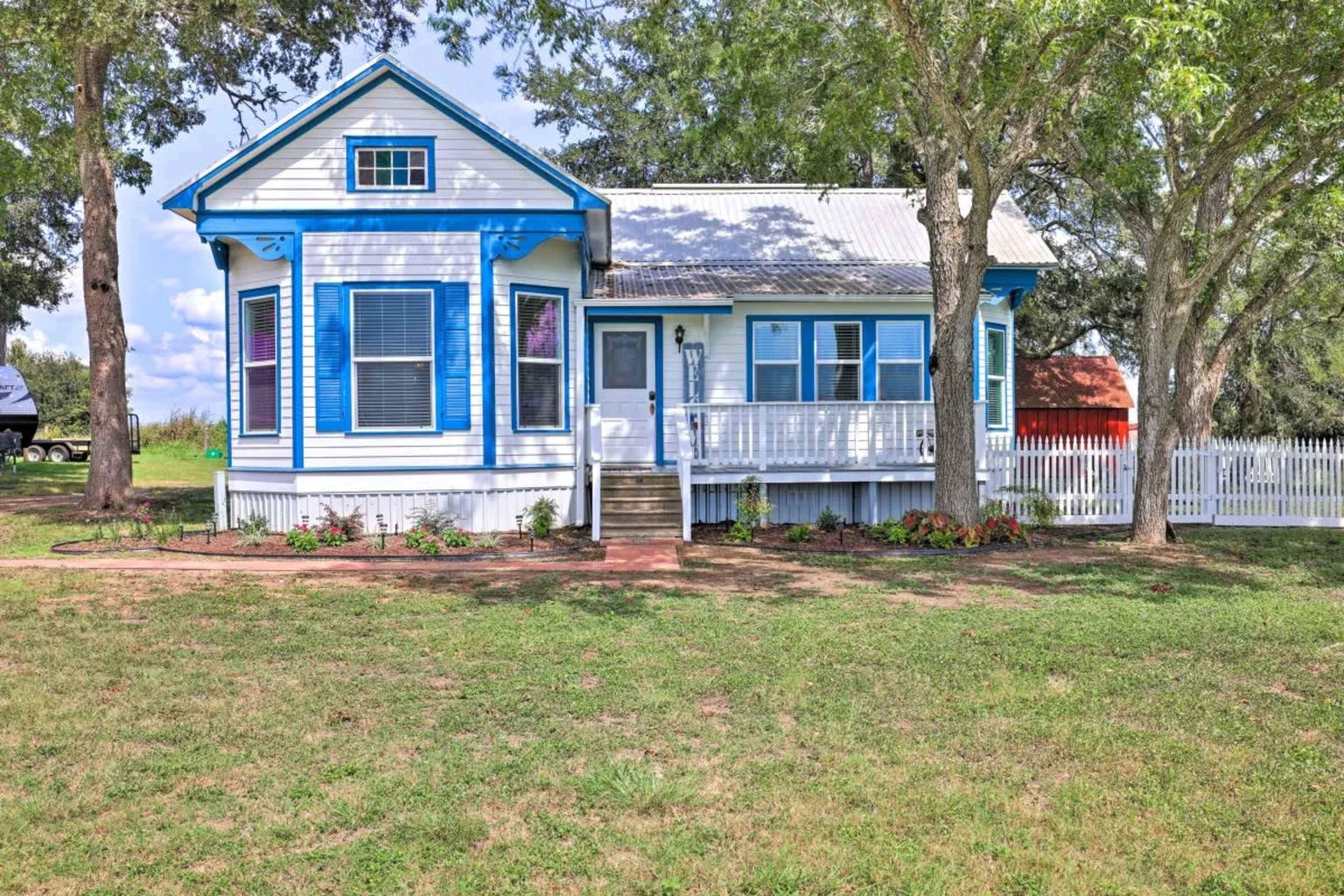 A white house with blue trim and a front porch is surrounded by green grass and trees, with a white picket fence in the background.