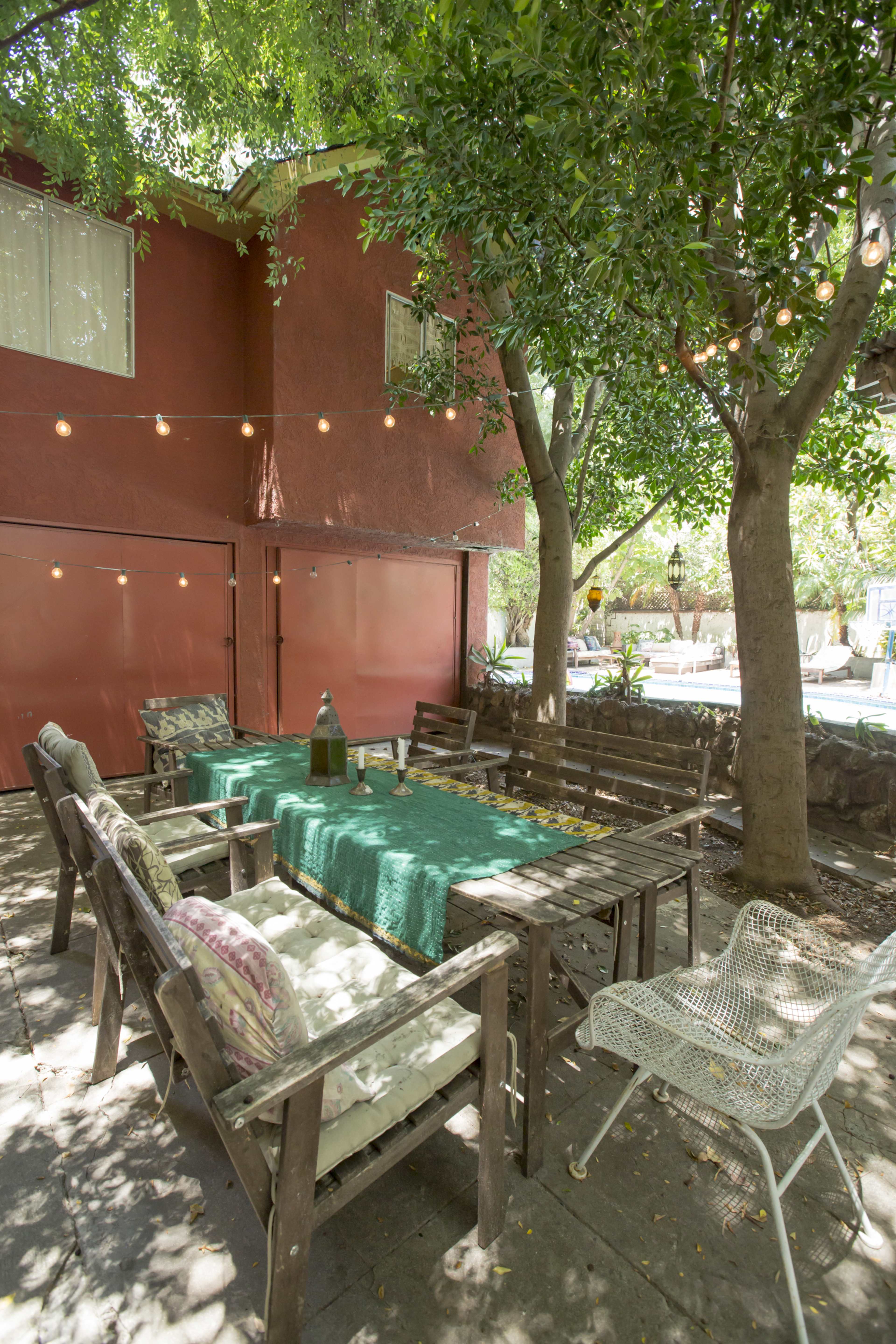 A wooden outdoor dining table is set under trees with string lights, surrounded by chairs in a shaded area near a red building.