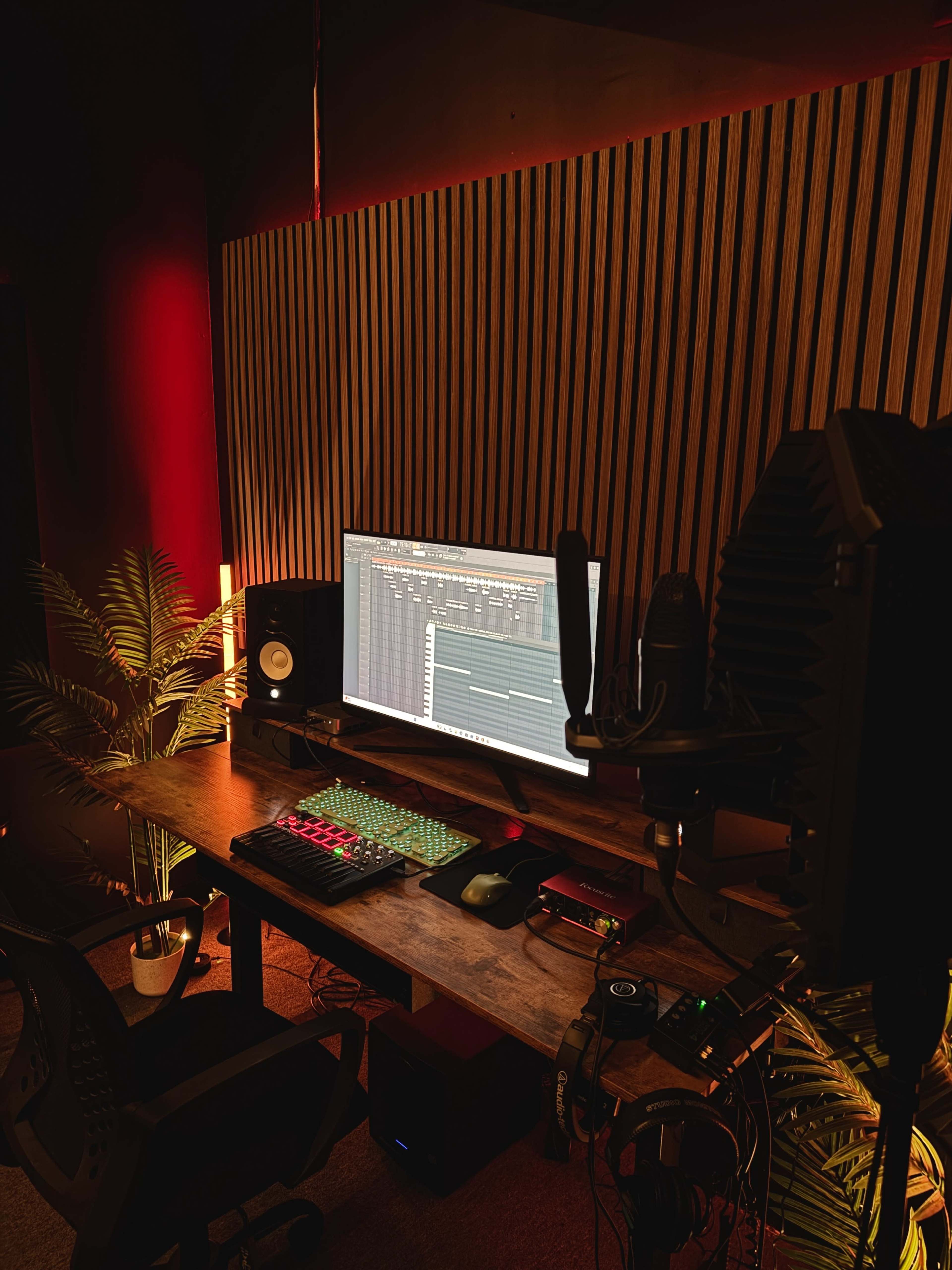 A music production studio setup features a computer monitor with editing software, a keyboard, studio monitors, and a microphone, surrounded by decorative plants and ambient lighting.