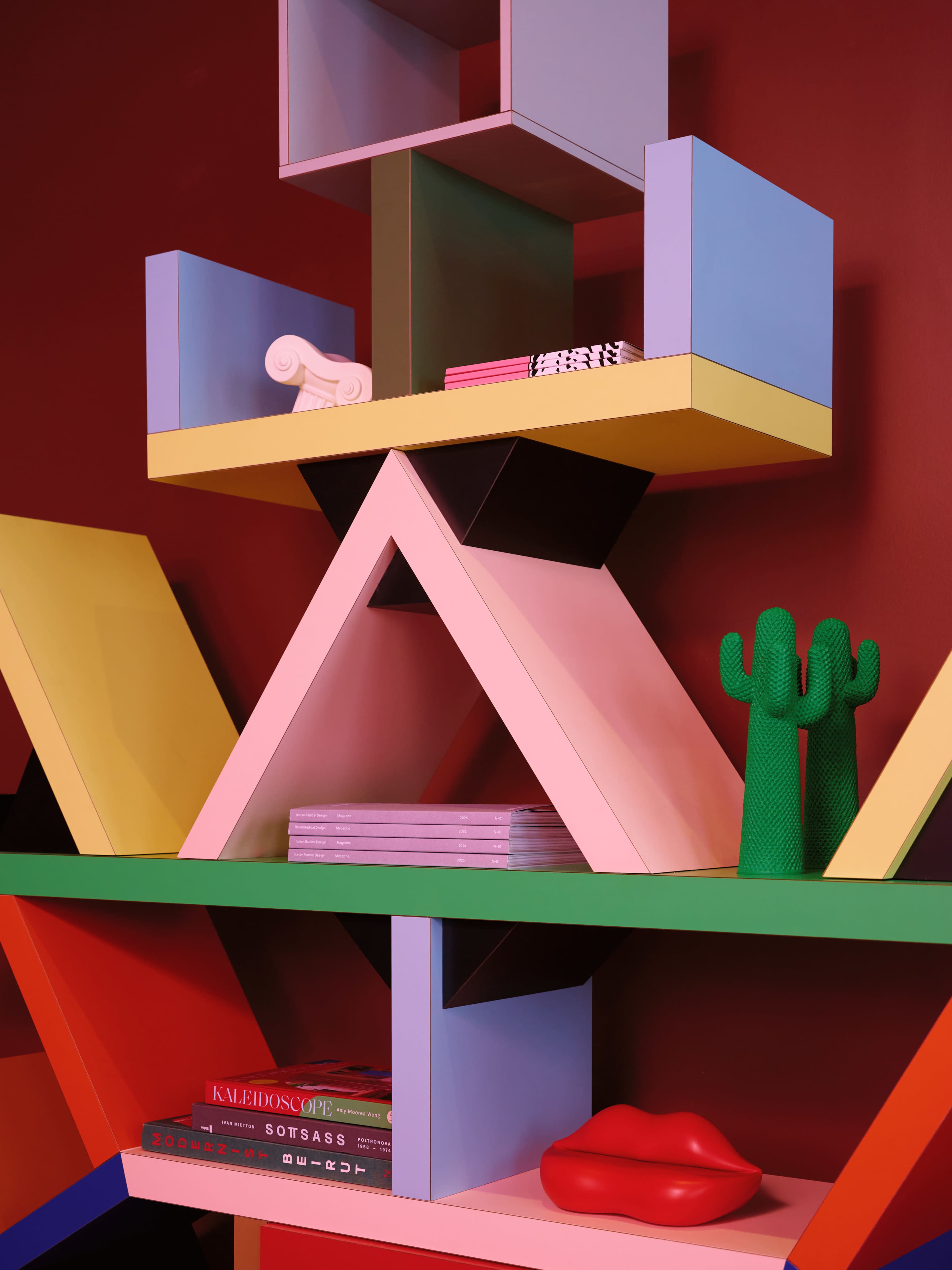 The image showcases a colorful geometric bookshelf displaying various decorative items, including books, a cactus sculpture, and a glossy lip-shaped object against a red wall.