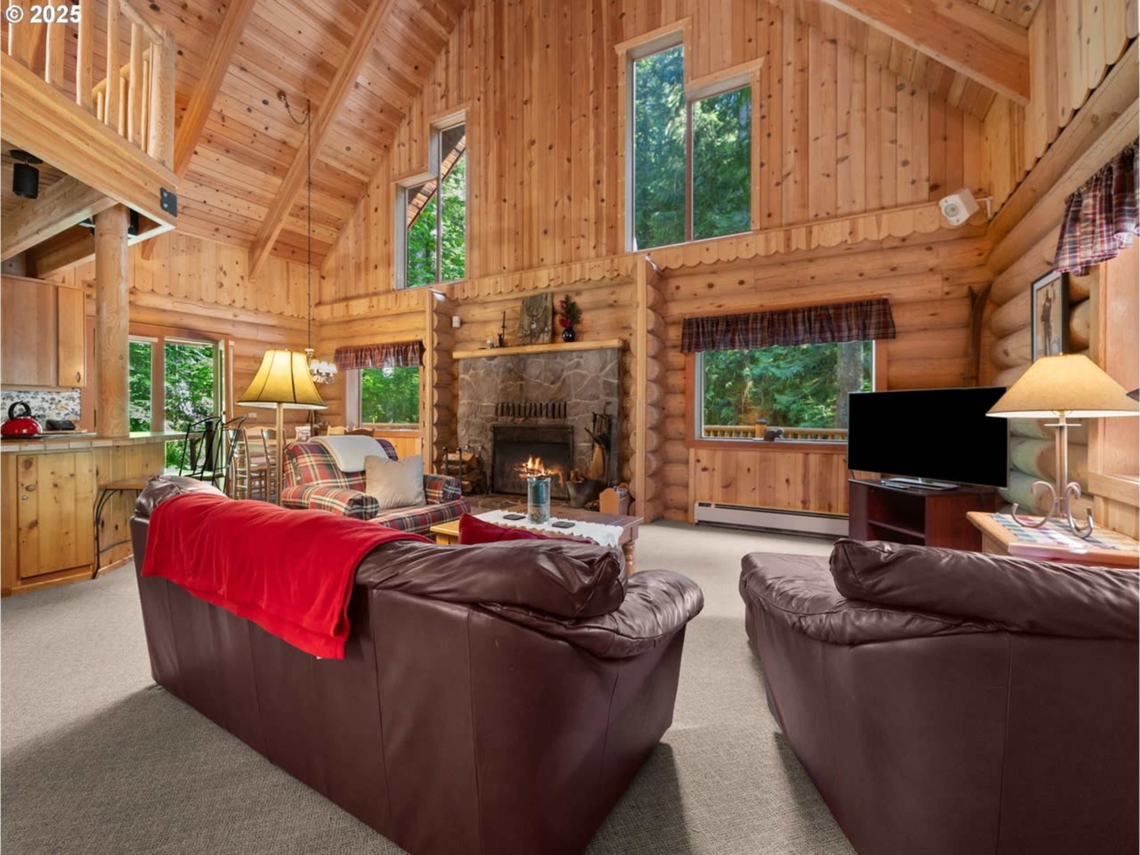 The image depicts a cozy log cabin interior with a stone fireplace, wooden beams, and large windows allowing natural light in.