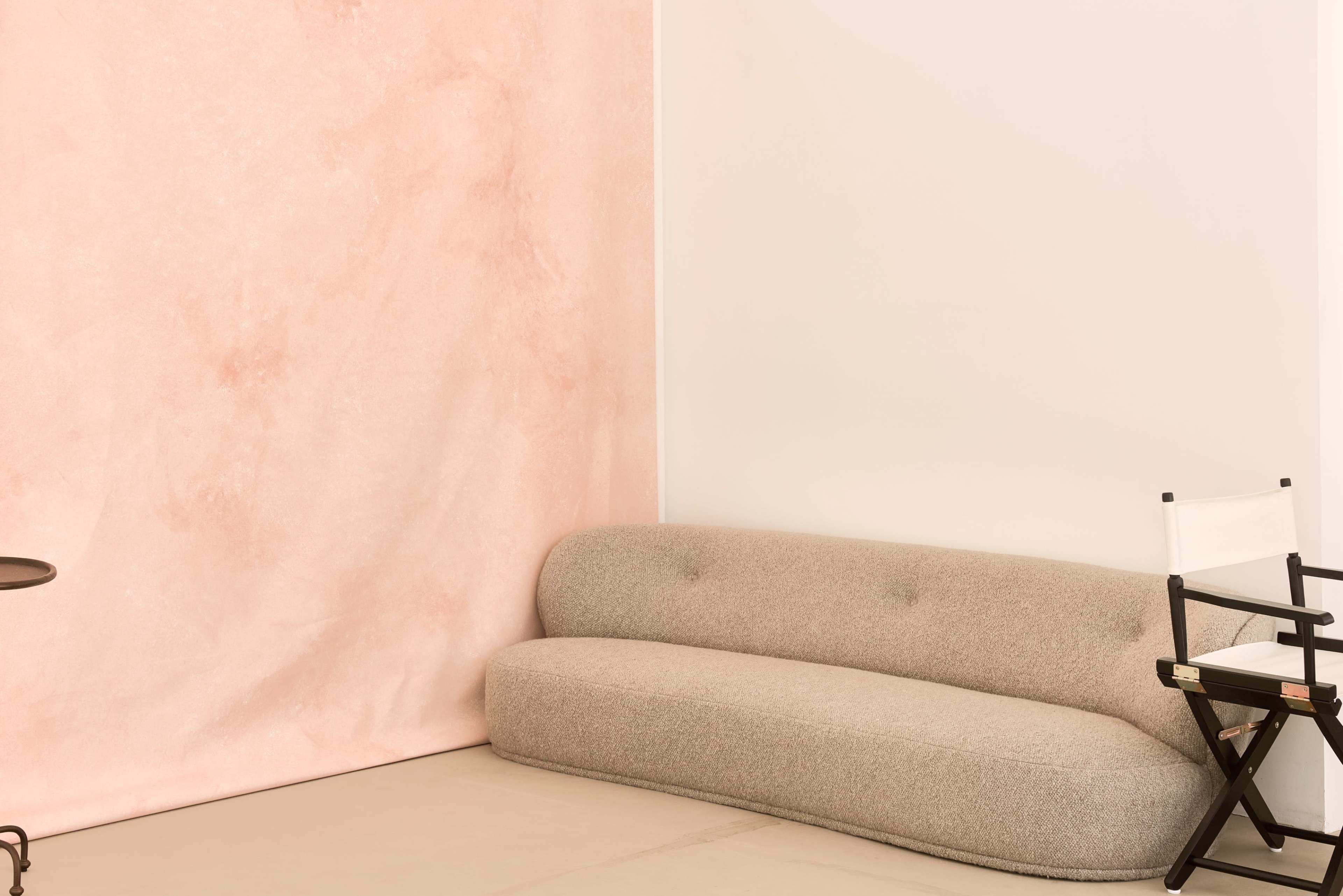 The image shows a minimalist interior featuring a curved beige sofa, a black and white director's chair, and a textured pink wall.