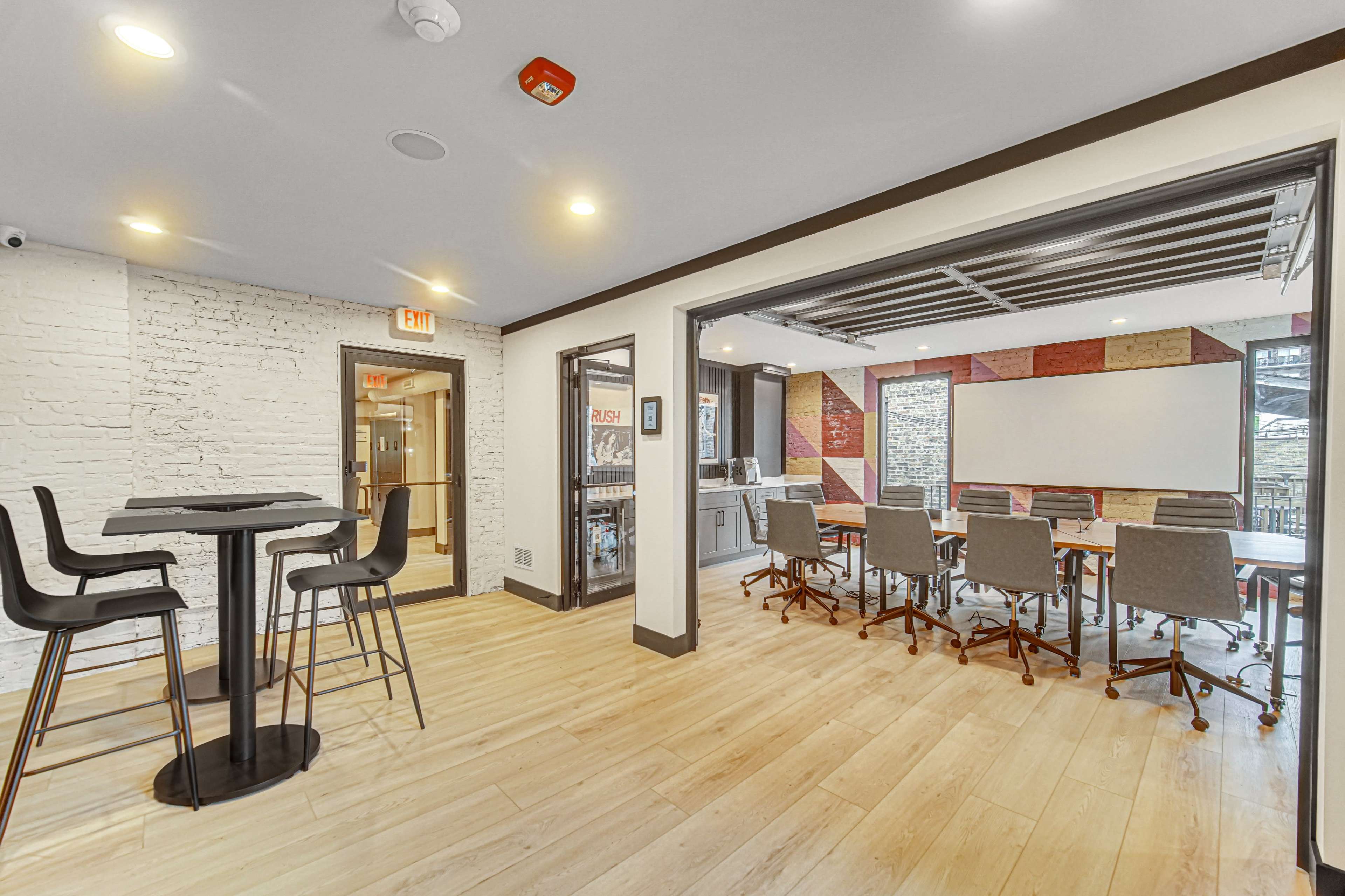 The image shows a modern office space featuring a meeting room with a large screen and a seating area with high tables and chairs.