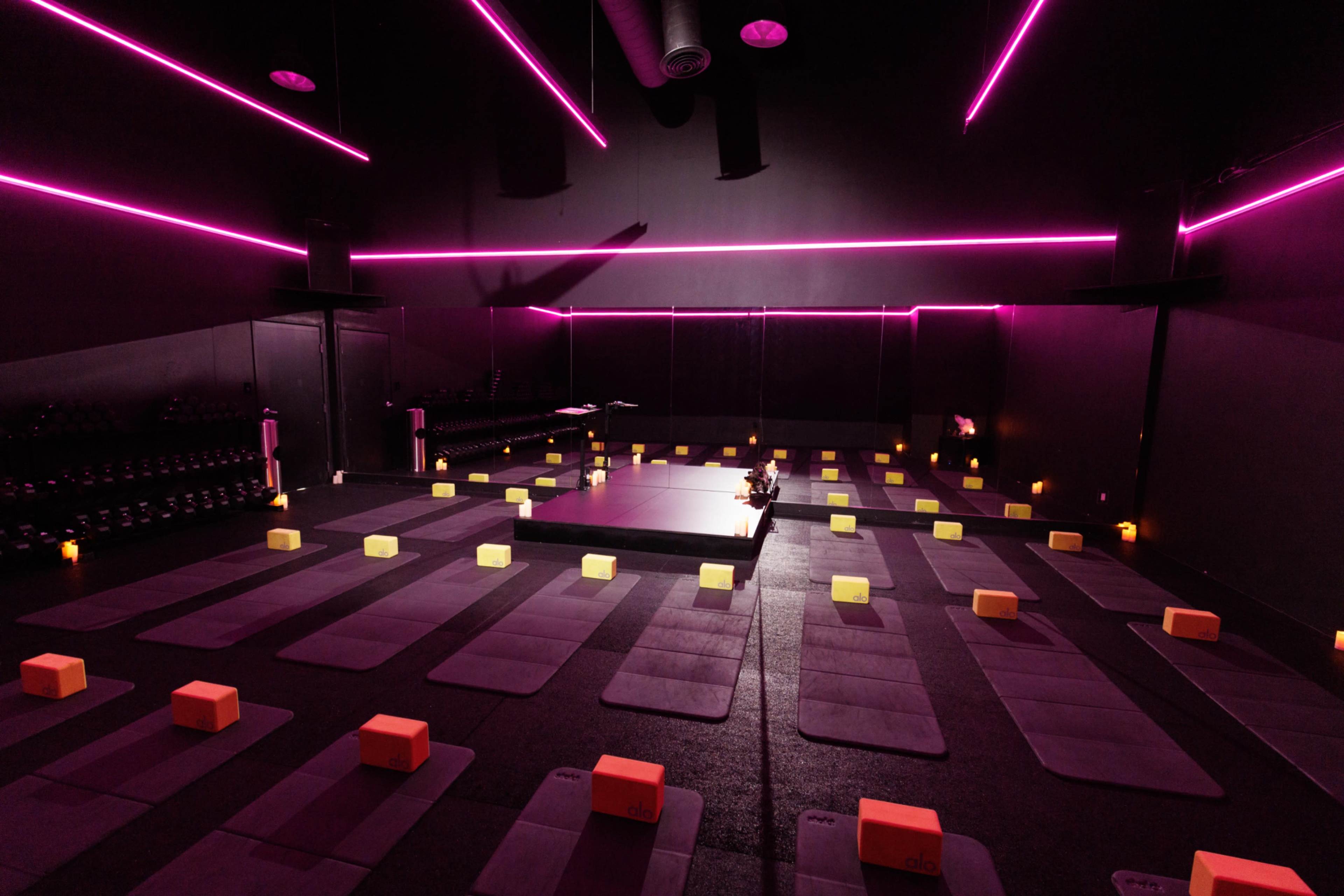 A dimly lit yoga studio features purple mats arranged neatly in rows, surrounded by orange yoga blocks and illuminated by pink accent lighting.