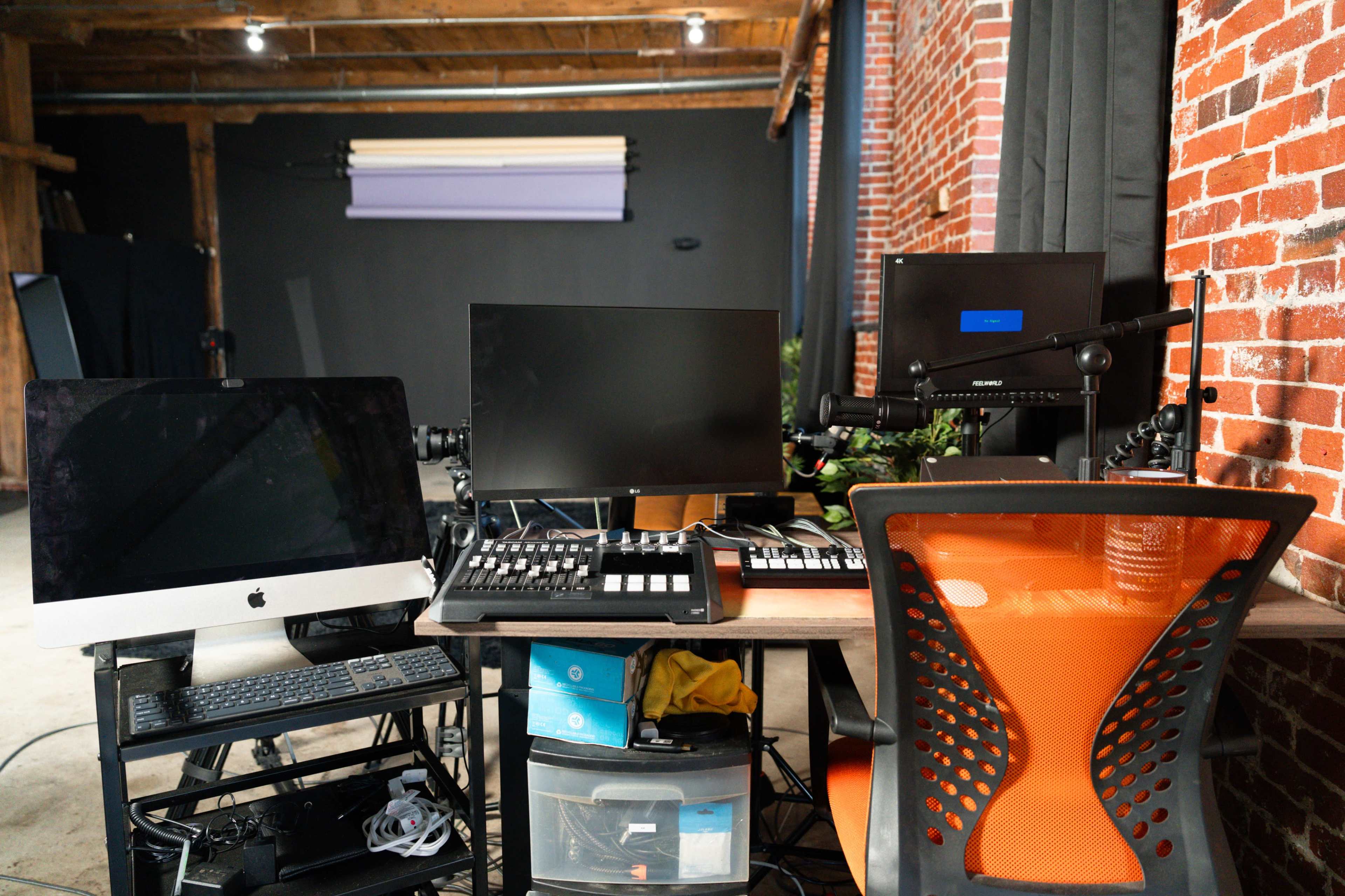 A workspace features multiple computer monitors, a keyboard controller, and an orange office chair set against a brick wall backdrop.