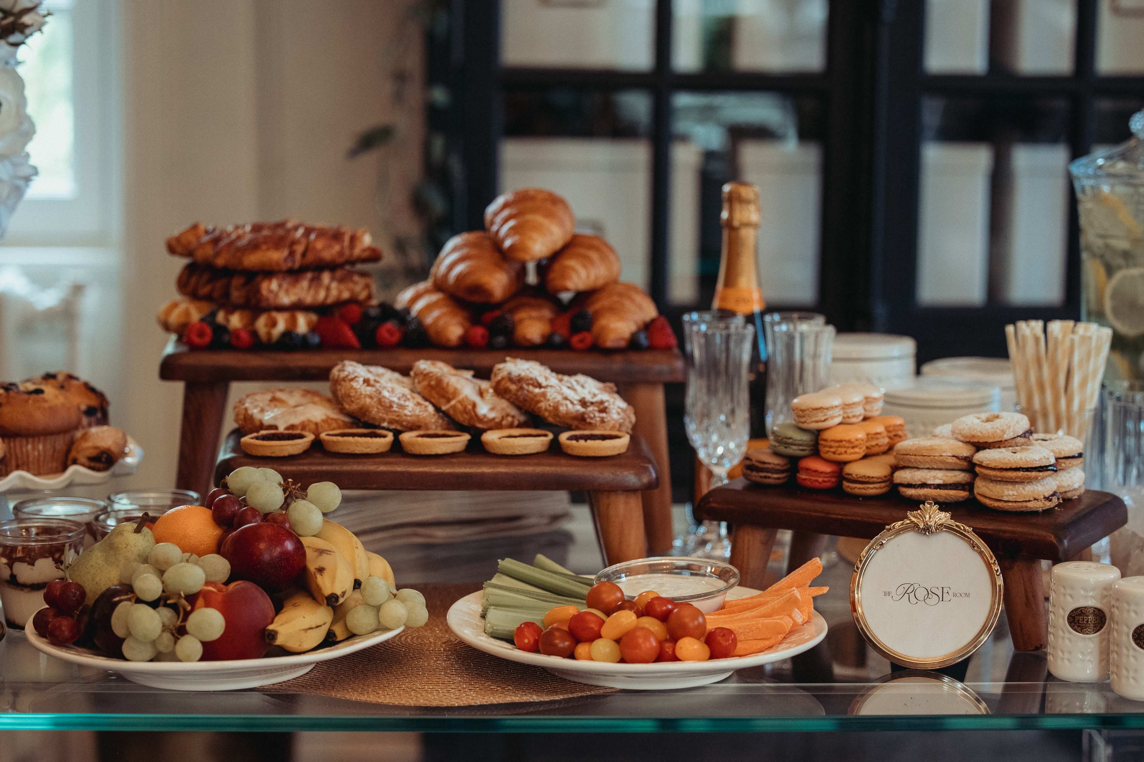 The image shows a neatly arranged food display featuring various baked goods, fruits, vegetables, and pastries on wooden stands and plates.