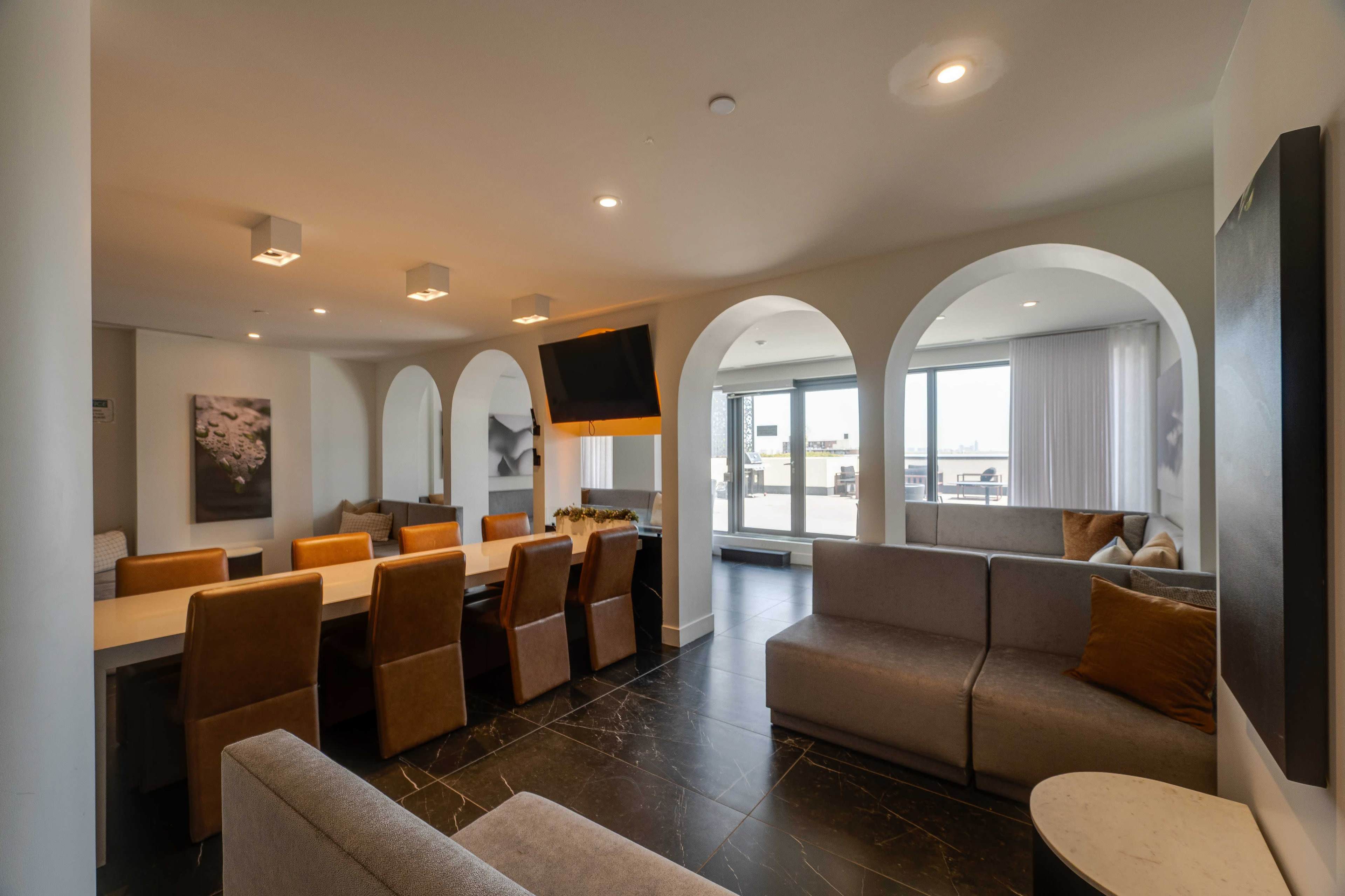 The image shows a spacious modern lounge area with arched doorways, a long dining table, gray sofas, and large windows that provide a view of the outdoors.
