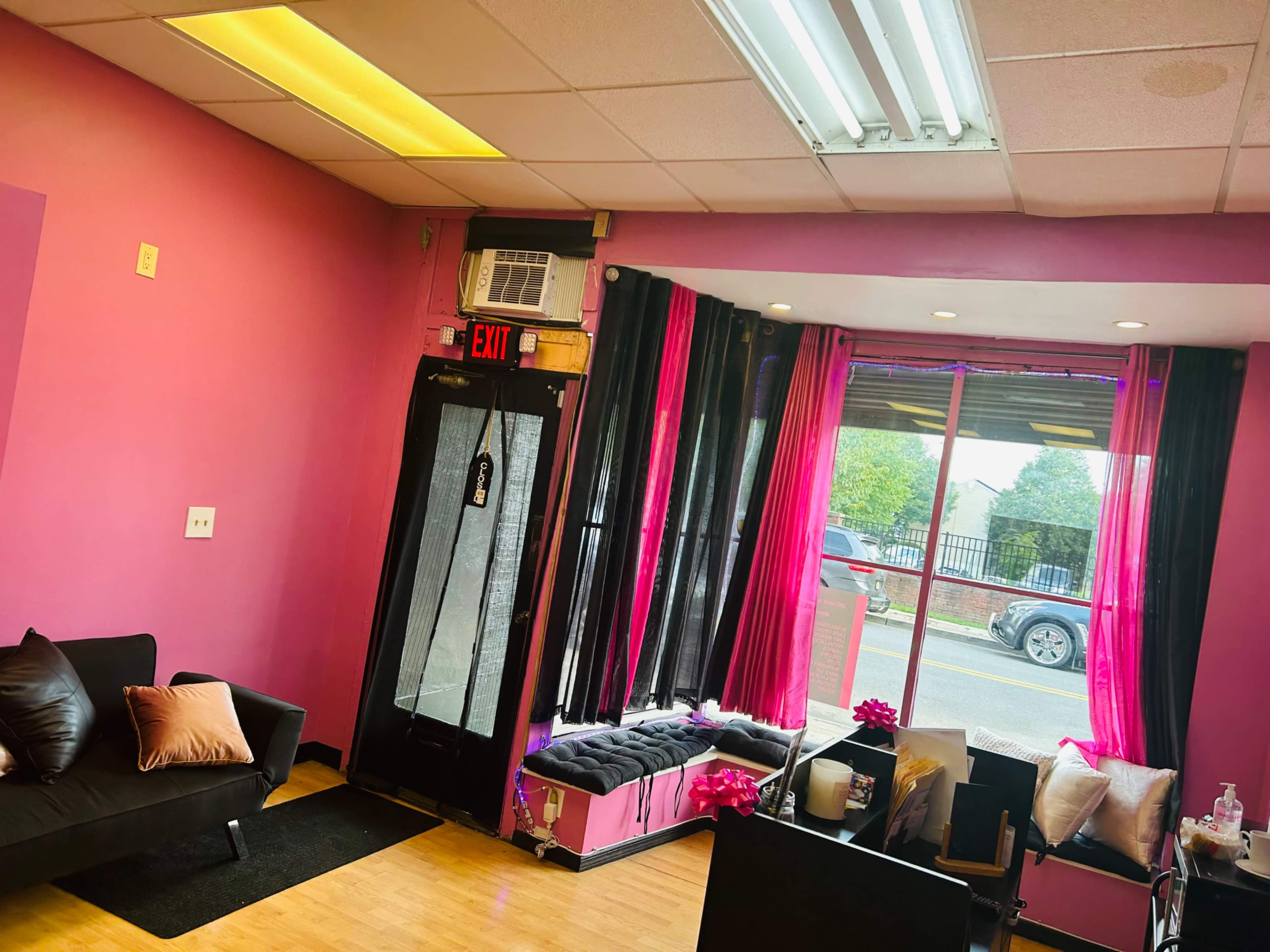 The interior of a salon features pink walls, a black sofa, and large windows with sheer curtains, along with a view of a parked car outside.