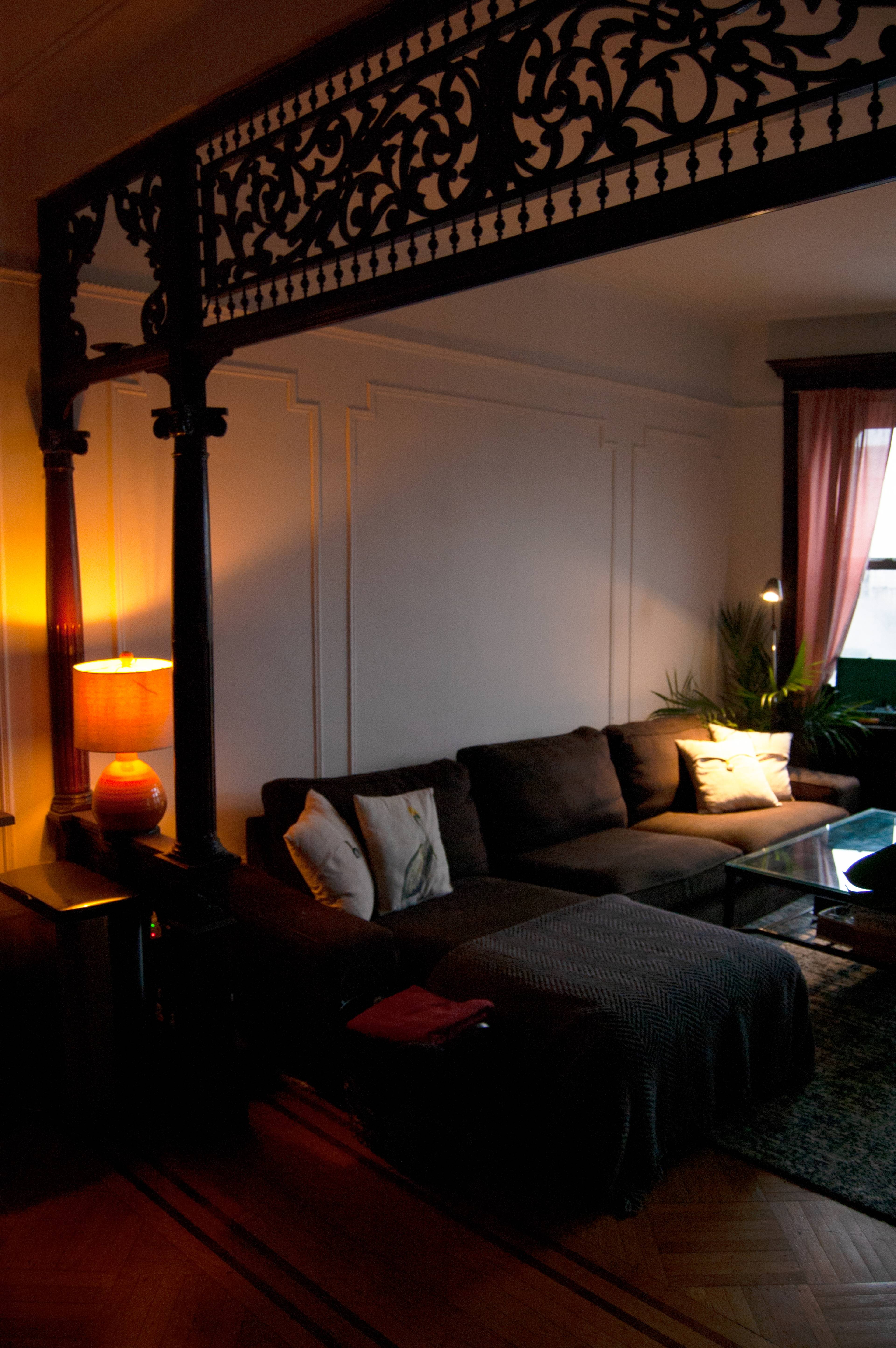 A cozy living room features a sectional sofa with decorative pillows, a glass coffee table, and a lamp casting soft light, framed by an ornate architectural divider.
