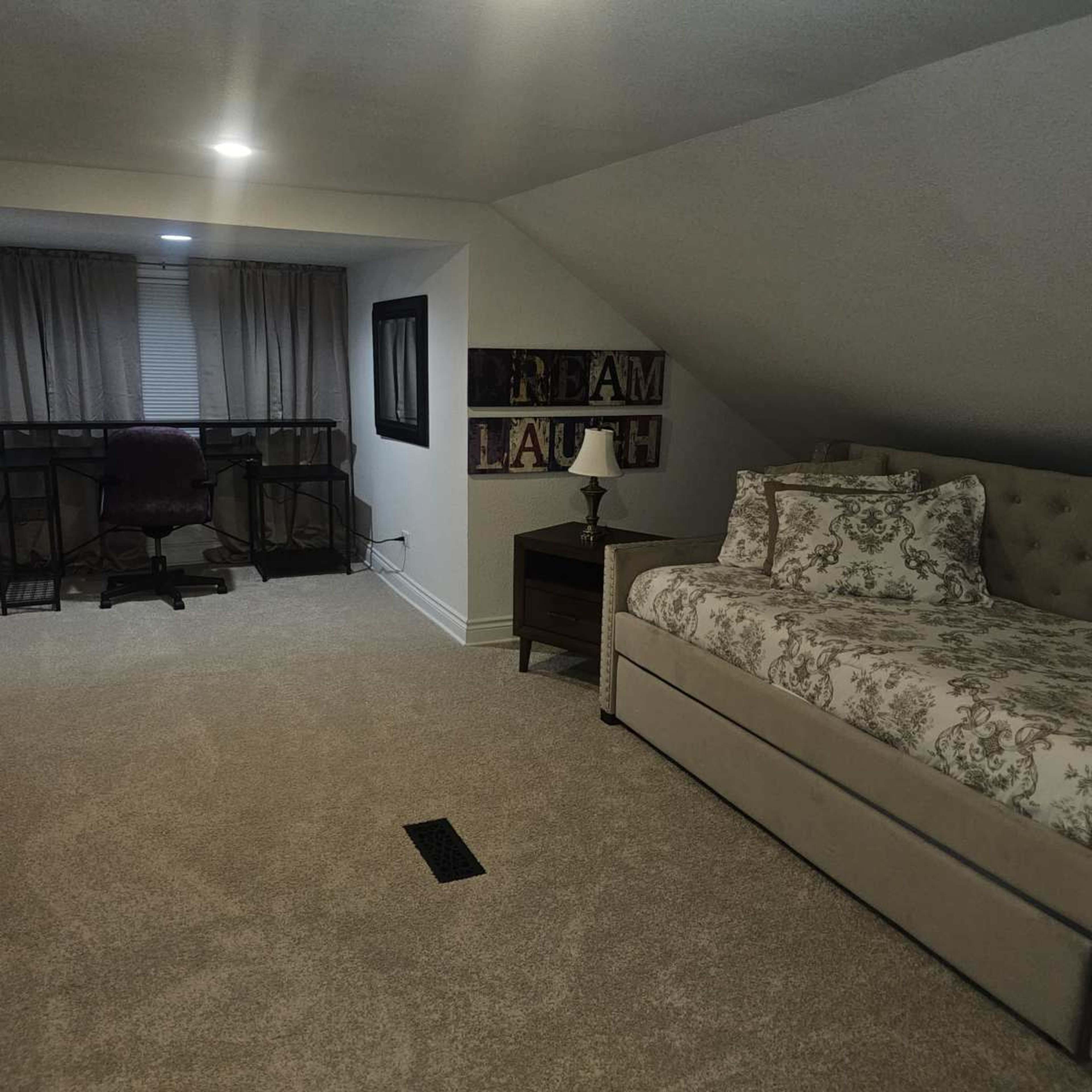 The image shows a bedroom with a single bed, a small nightstand with a lamp, a desk with a chair, and large windows covered with curtains.