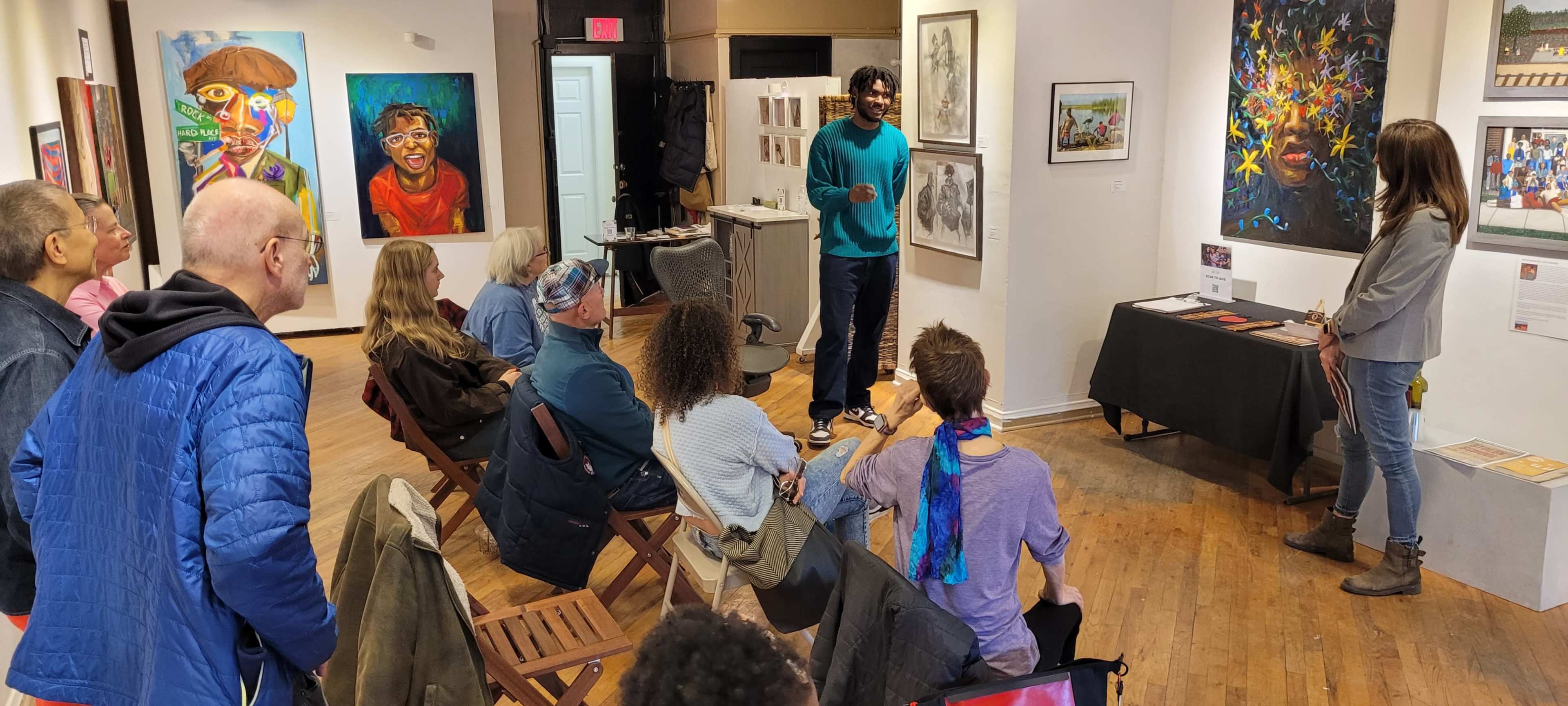 A diverse group of people is gathered in an art gallery where a presenter is discussing artwork, while various pieces hang on the walls.