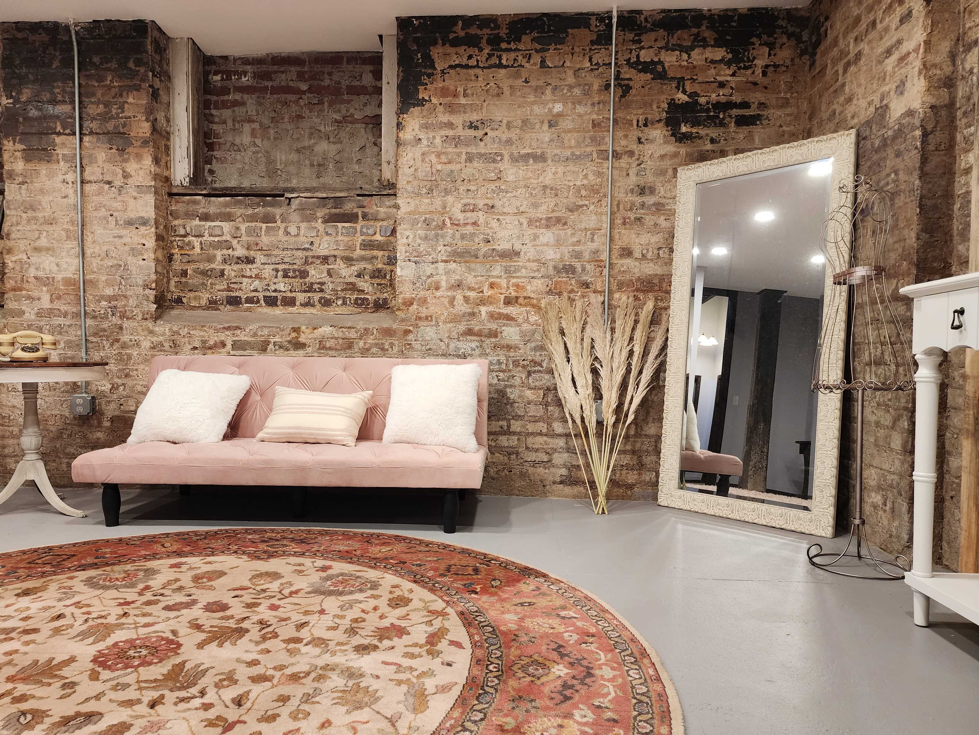 A cozy room featuring a pink sofa with cushions, a large mirror, and a decorative rug against a backdrop of exposed brick walls.