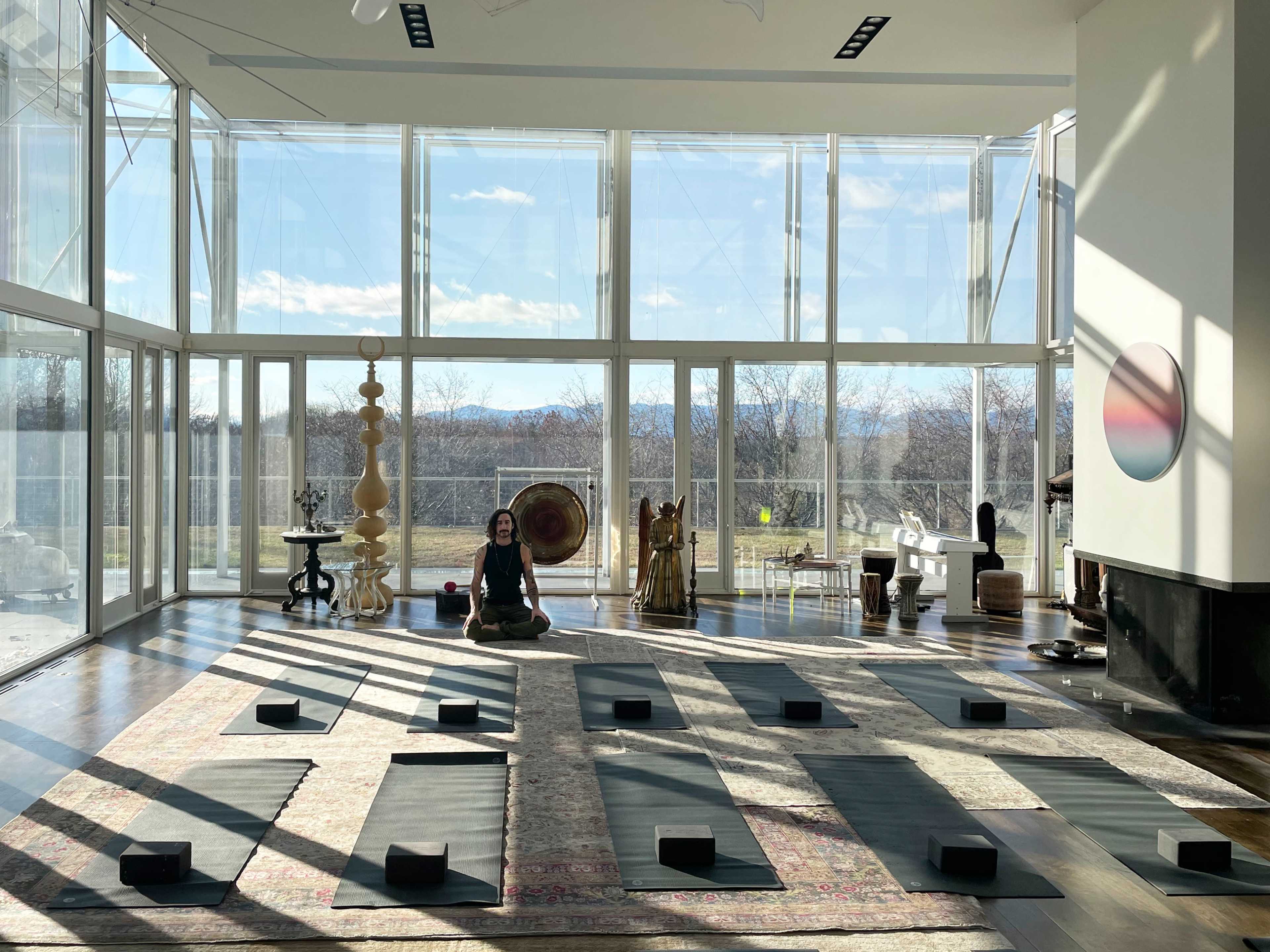 A spacious yoga studio with large windows, featuring yoga mats arranged in rows and a woman meditating at the front.