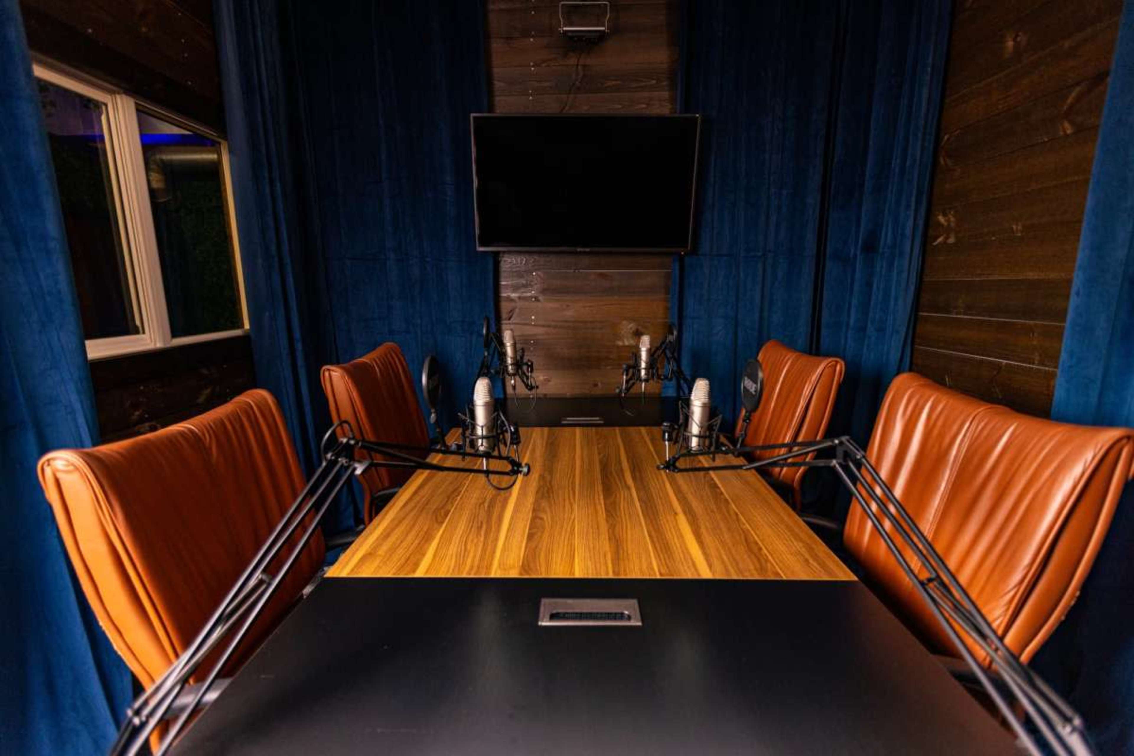 The image shows a recording studio with a table, four brown leather chairs, and two microphones positioned on either side, along with a television mounted on the wall.