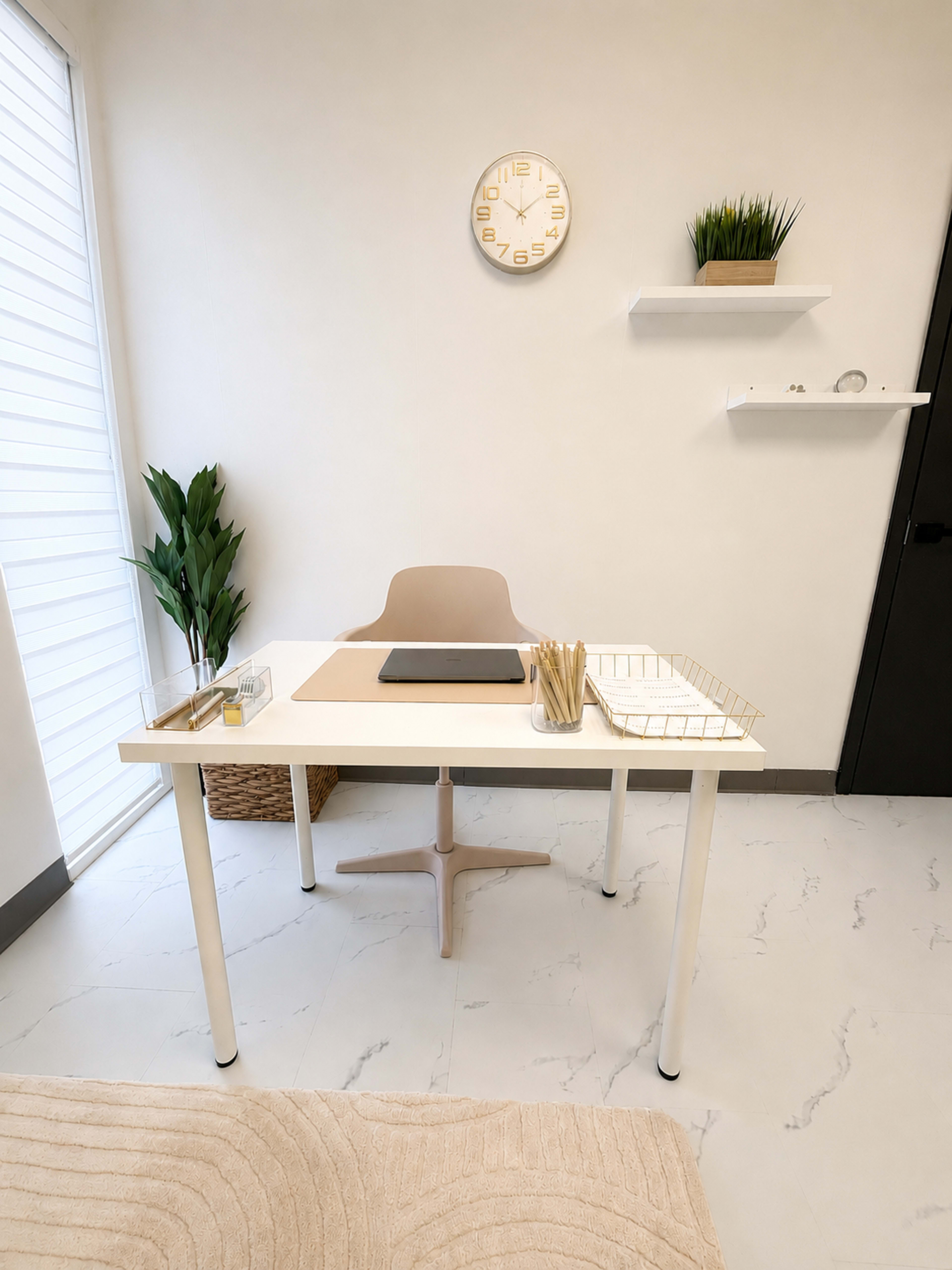 The image shows a minimalist office space featuring a white desk with a laptop, a few stationery items, a light-colored chair, and a plant, along with a clock on the wall and shelves with decorative items.