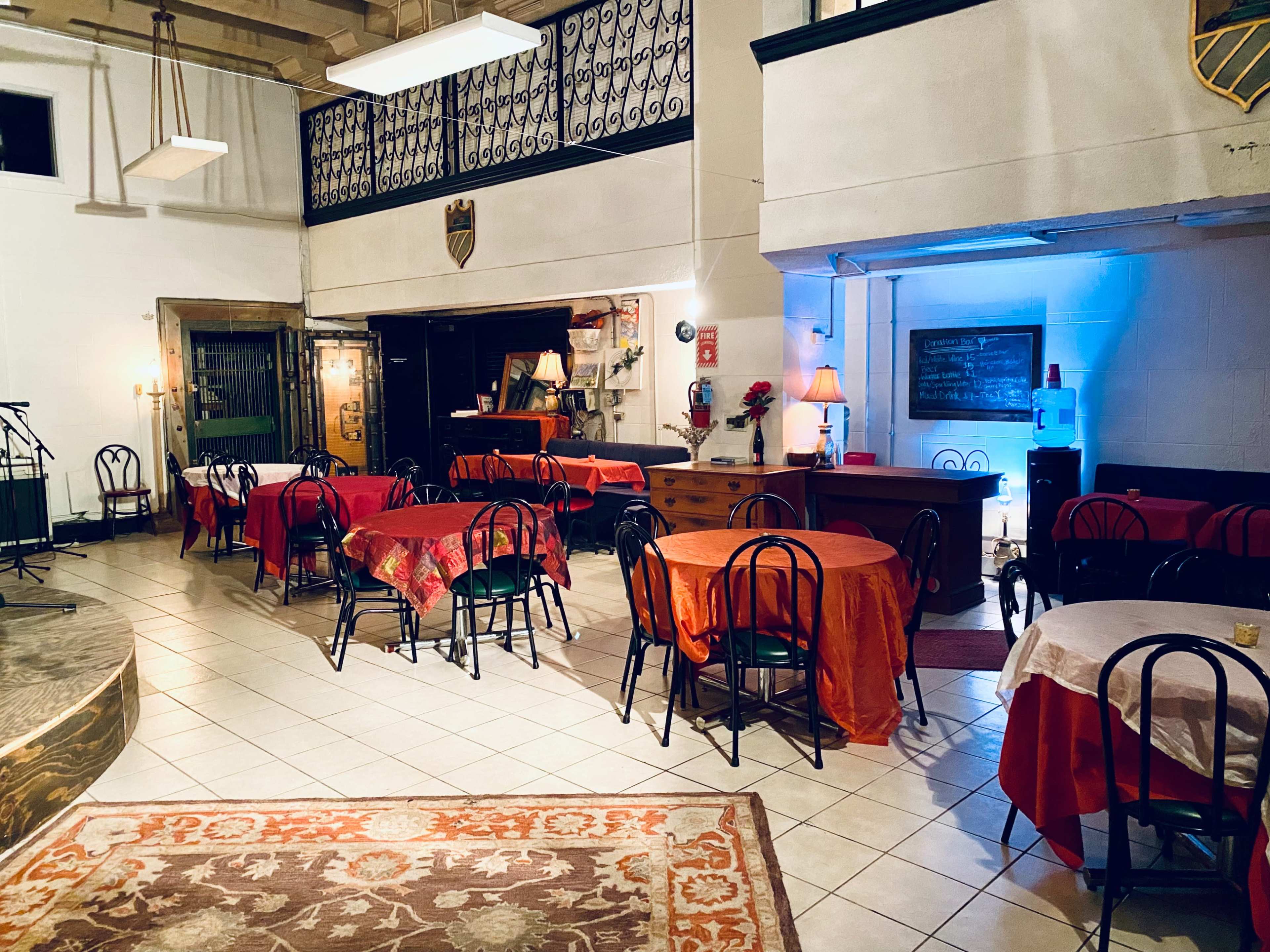 A dining area with several tables covered in red tablecloths, surrounded by black chairs, and features a small stage in the corner.