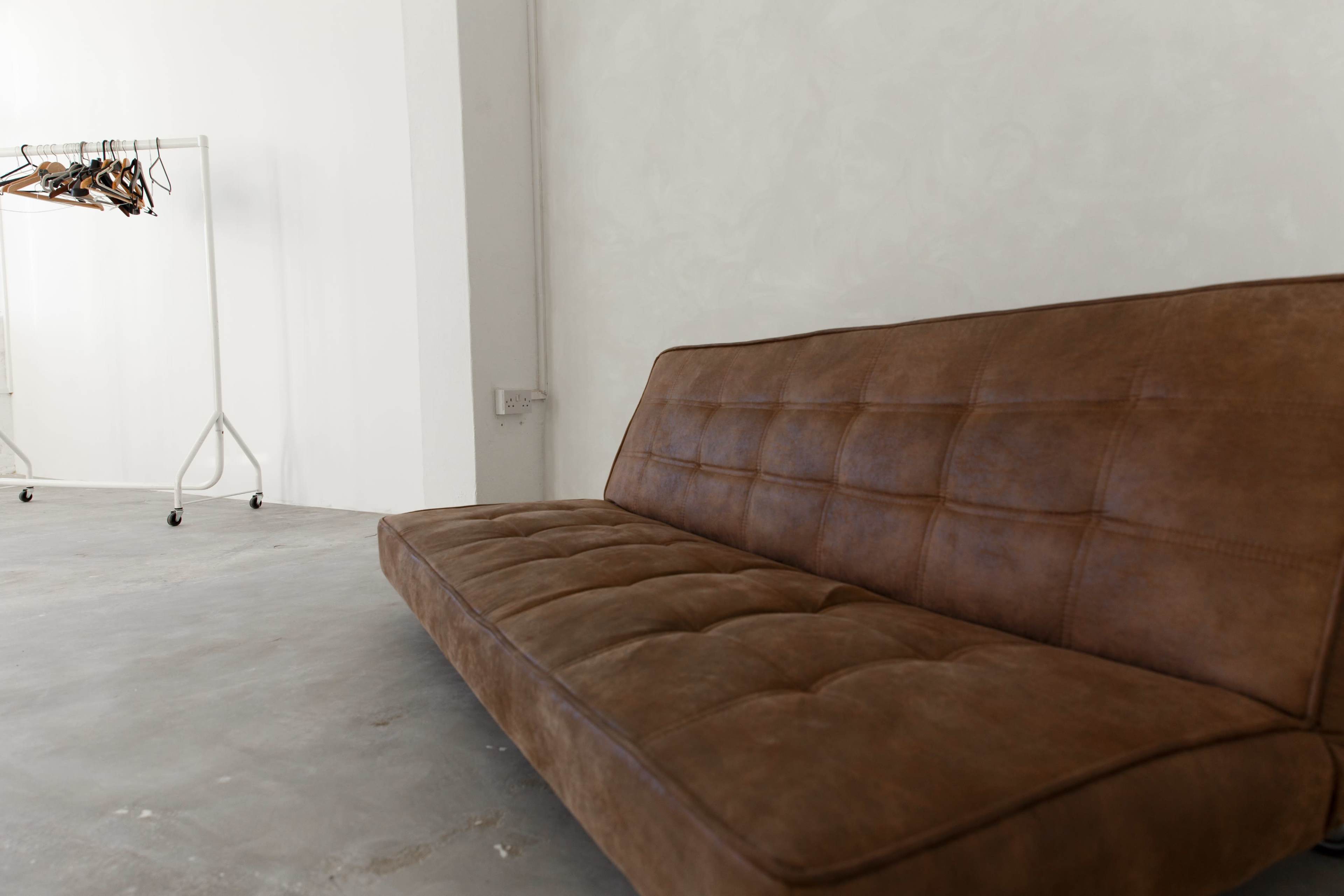 A brown leather sofa is positioned against a plain wall, with a clothing rack visible in the background.