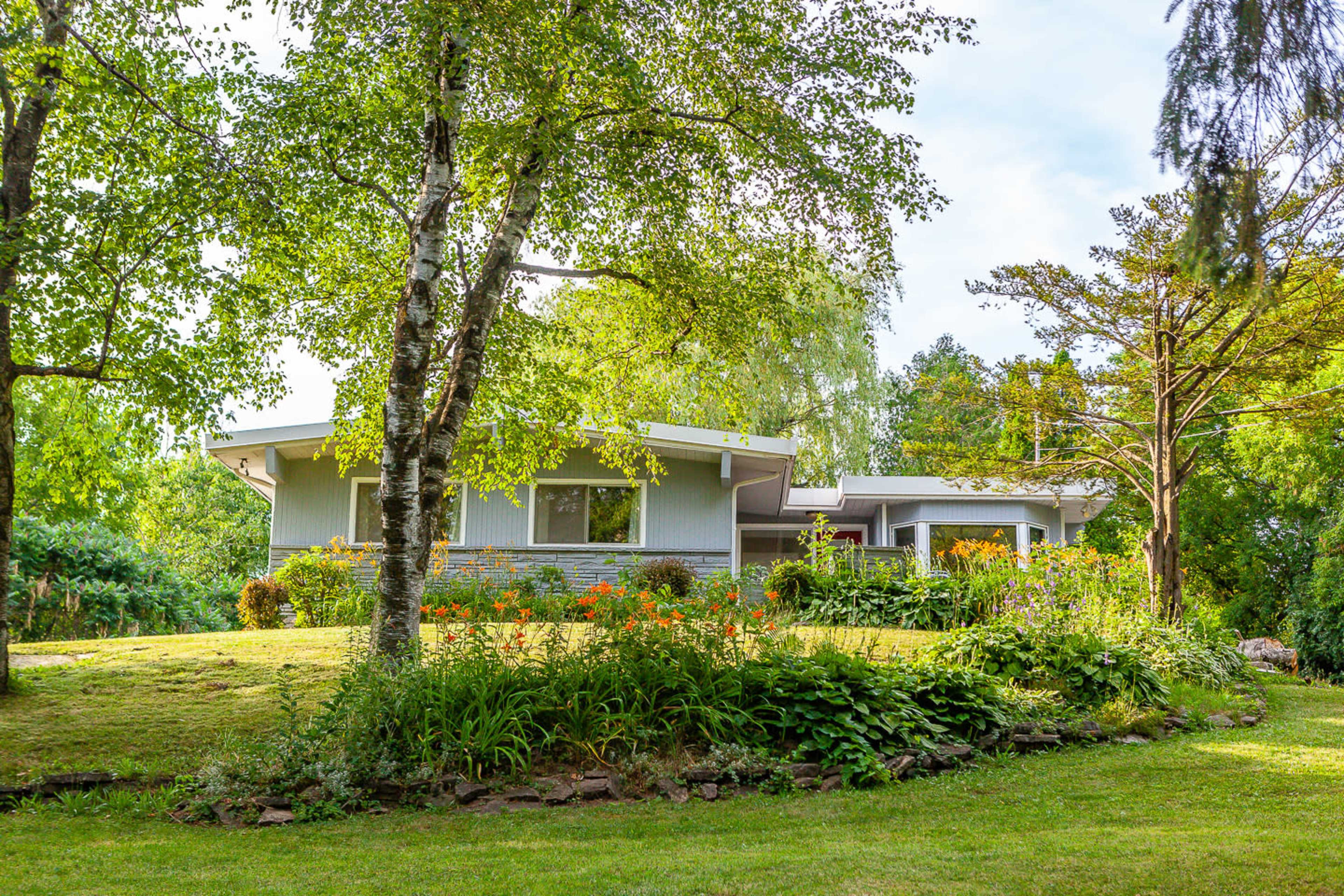 A modern house with large windows is surrounded by lush greenery and landscaped gardens, set against a clear blue sky.