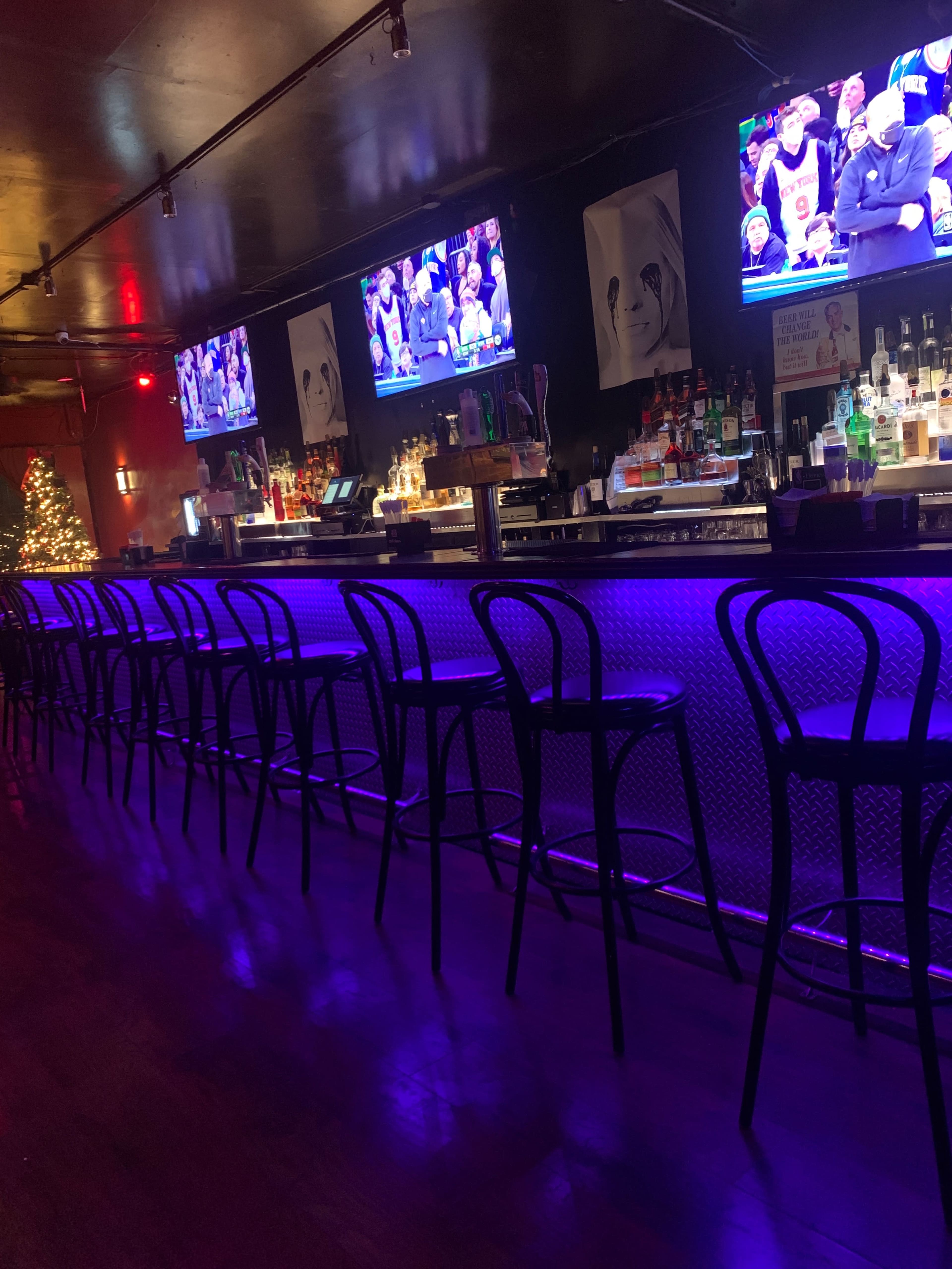 A dimly lit bar with several monitors displaying a basketball game, illuminated by blue LED lights along the counter.