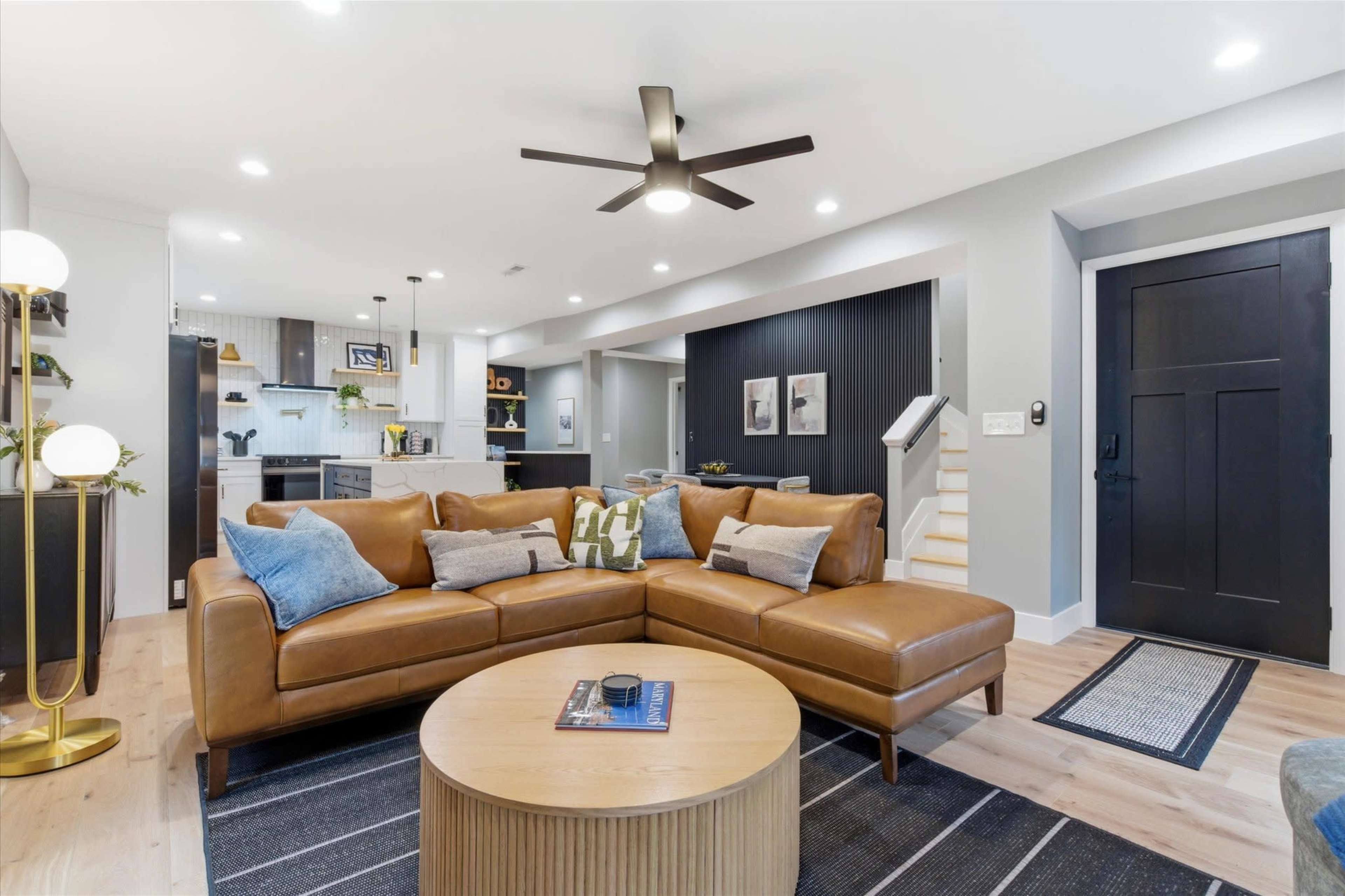The image shows a modern living room featuring a brown leather sectional sofa, a round coffee table, and a well-lit kitchen area in the background.