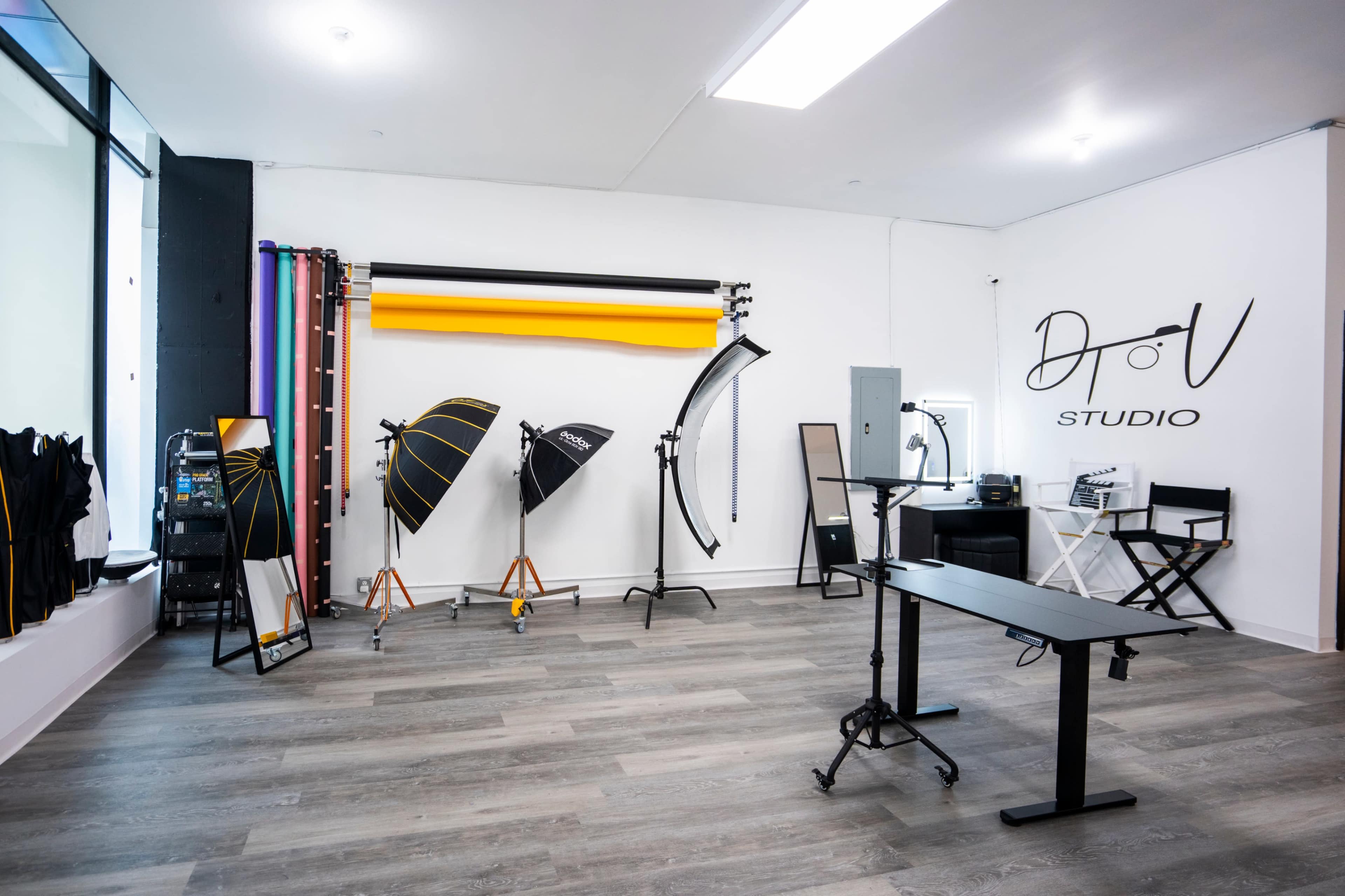 The image shows a photography studio equipped with various lighting setups, backdrops, and a workspace with chairs and equipment.