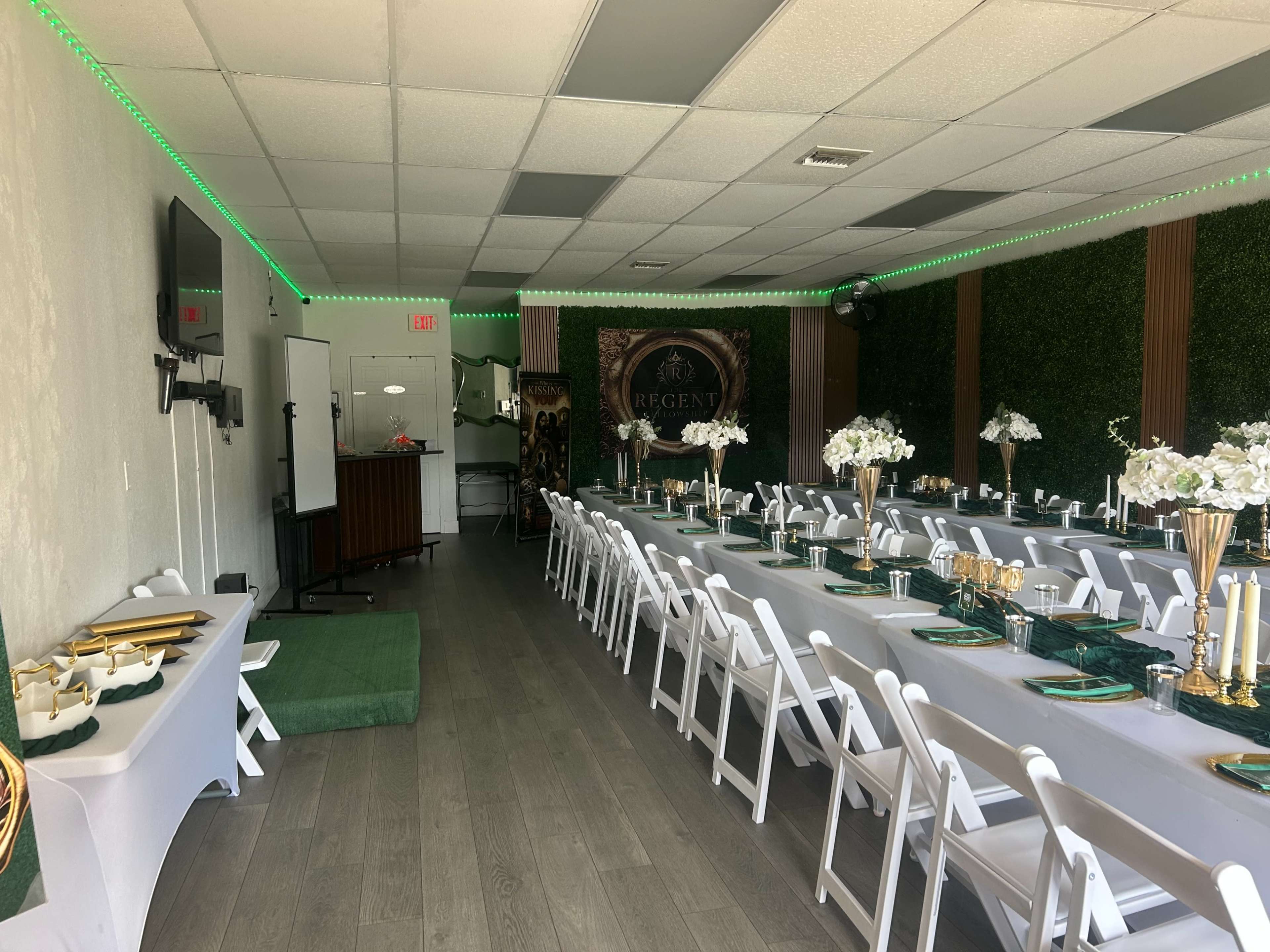 The image shows a banquet hall arranged with multiple tables covered in white tablecloths and centered with floral arrangements, illuminated by green LED lighting.