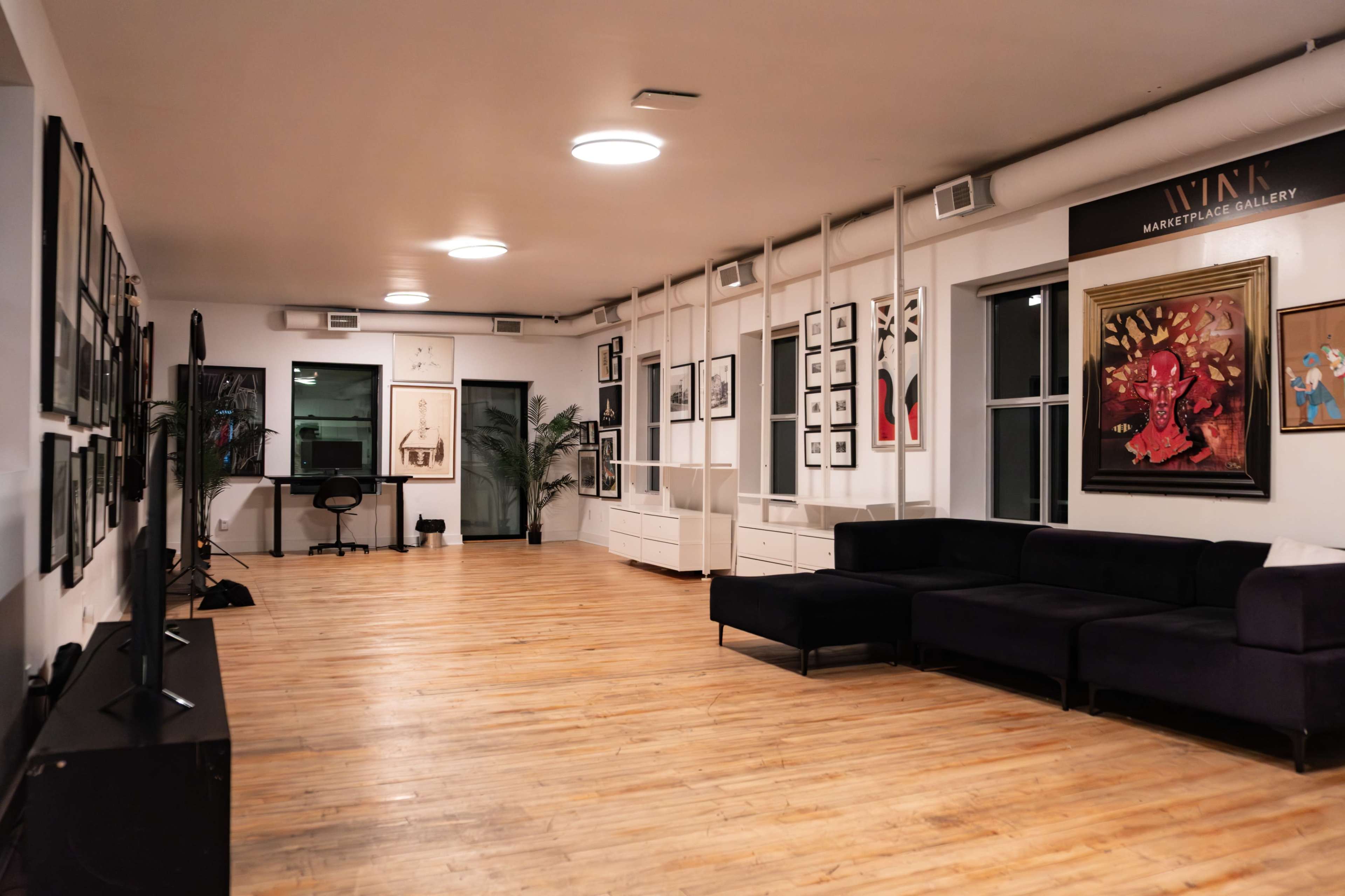 The image features an interior space of a gallery with wooden flooring, a dark sofa, framed artwork on the walls, and a sleek workstation in the background.