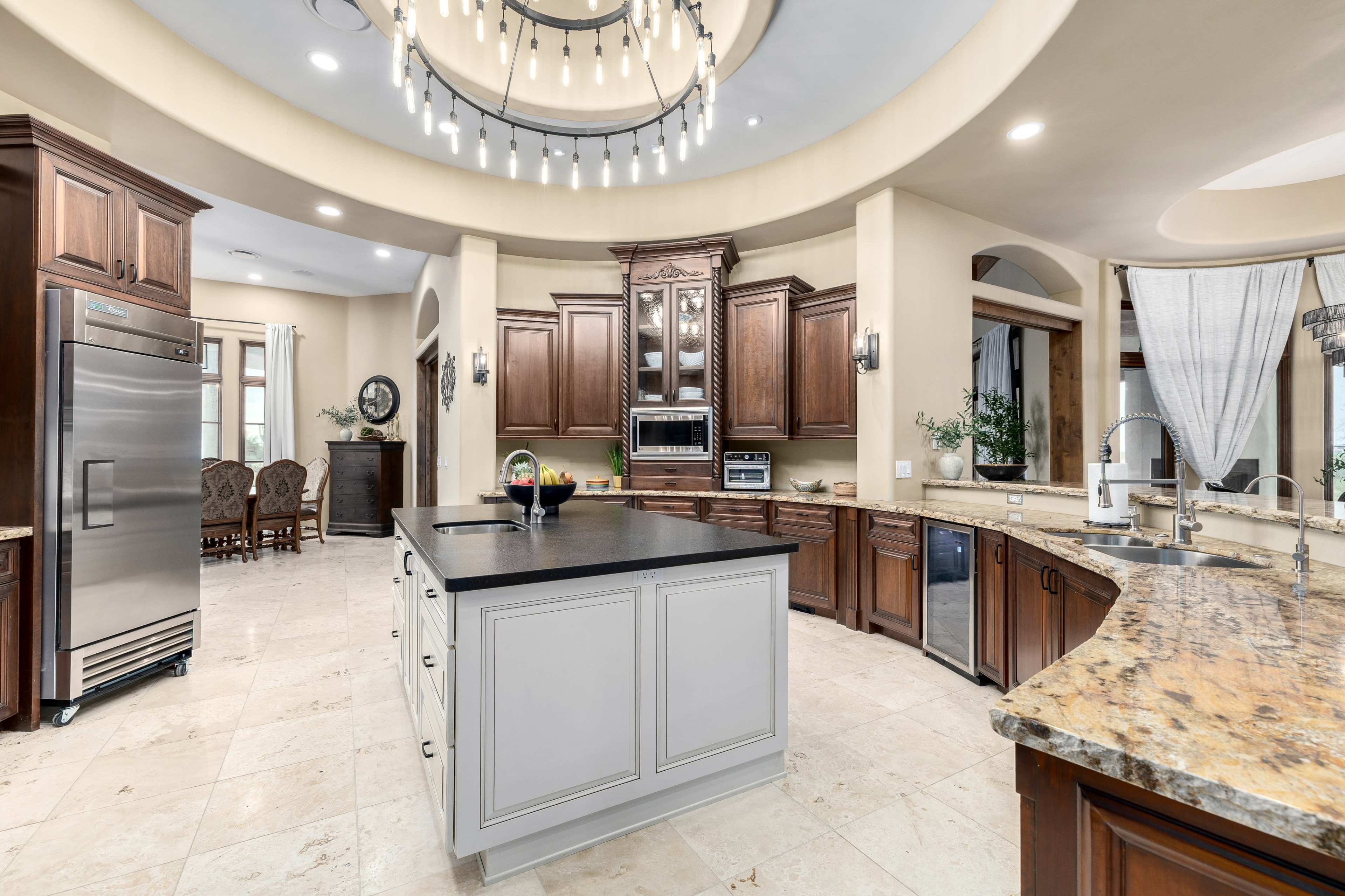 The image shows a spacious kitchen with an island, stainless steel appliances, and wooden cabinets, illuminated by a circular chandelier.