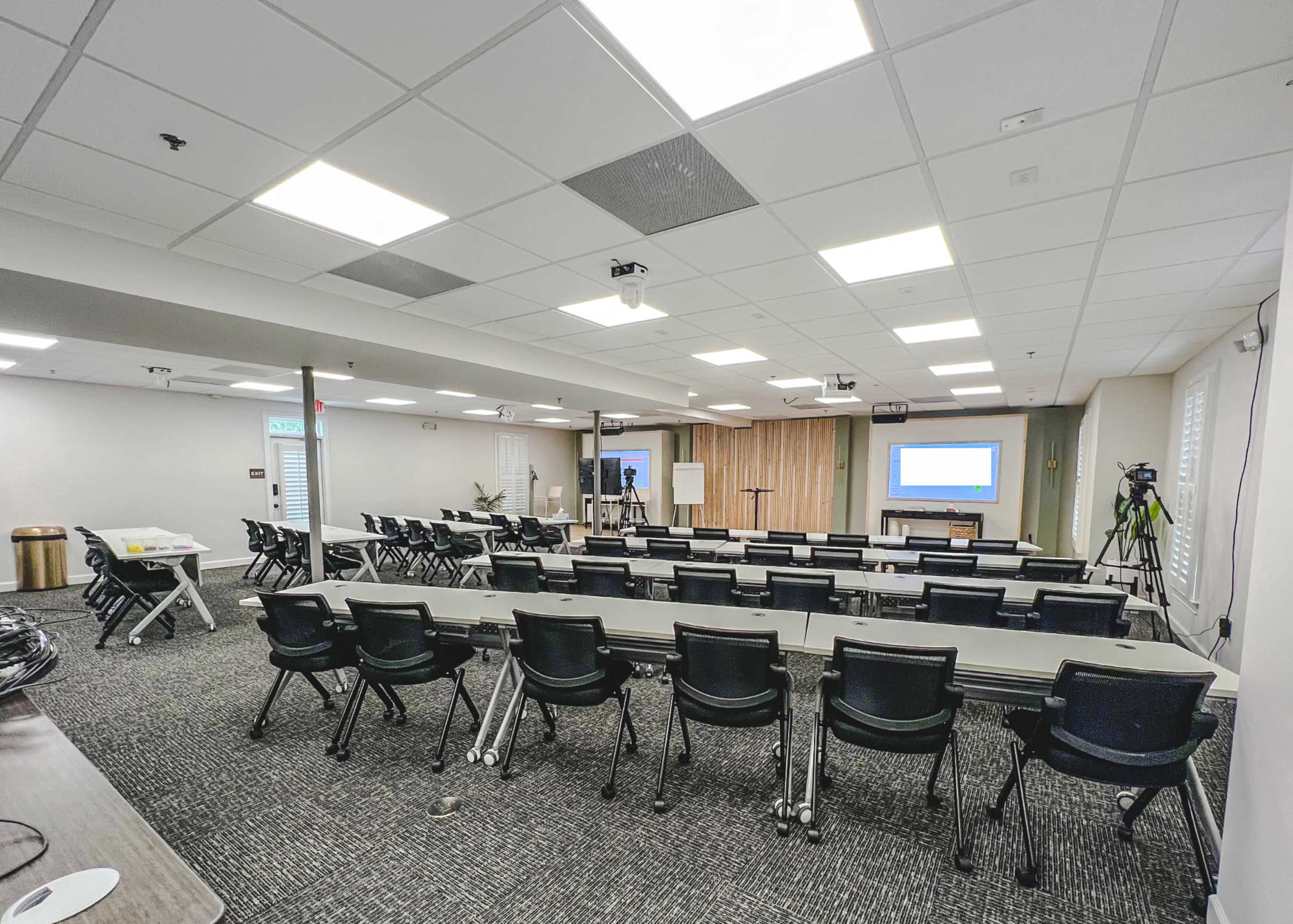 A conference room set up with rows of chairs facing two presentation screens at the front, with additional equipment for recording or broadcasting.