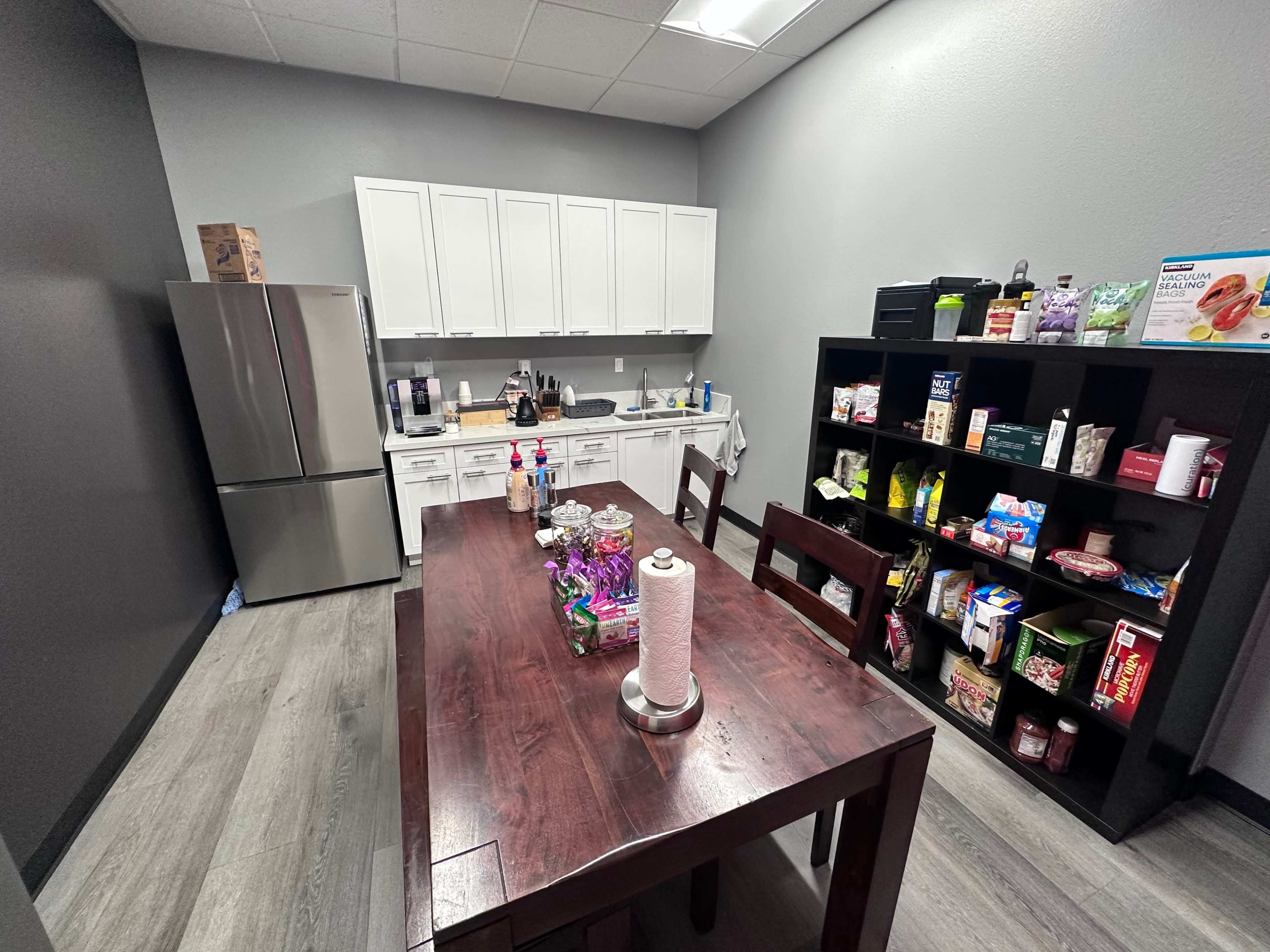 A kitchen area with a dining table, a refrigerator, white cabinets, and a shelf filled with various food items and supplies.