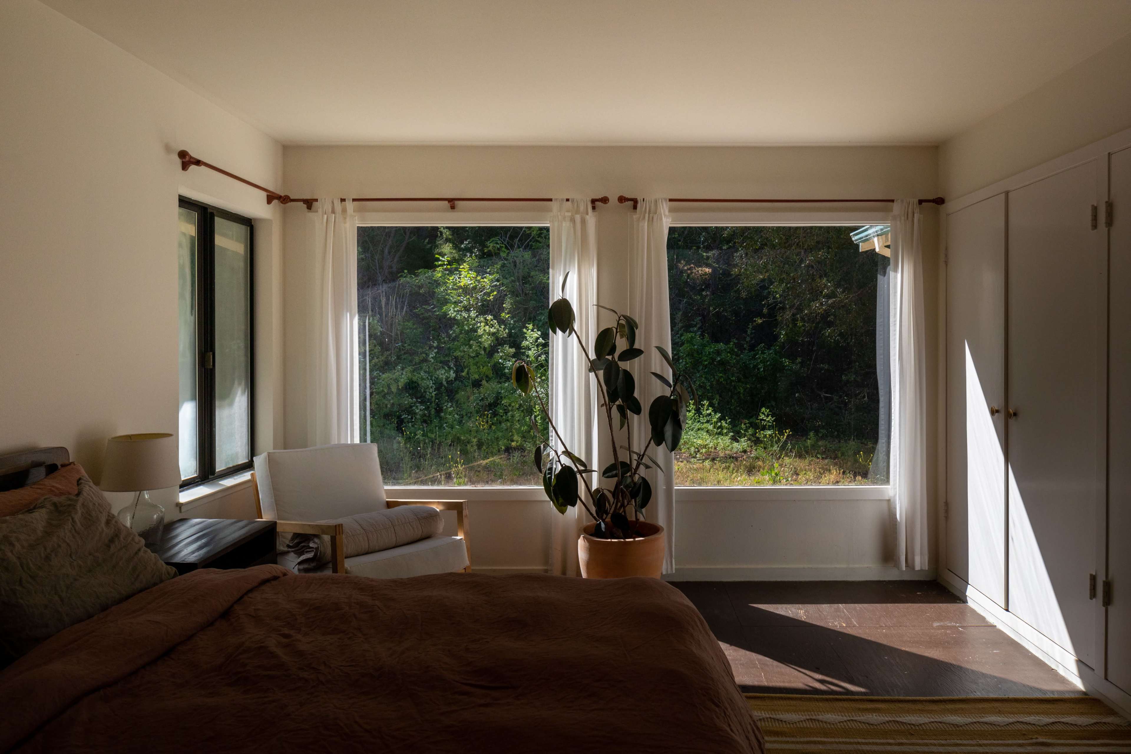A bright bedroom features large windows with sheer curtains, a bed with a brown cover, a chair next to a plant, and a view of greenery outside.