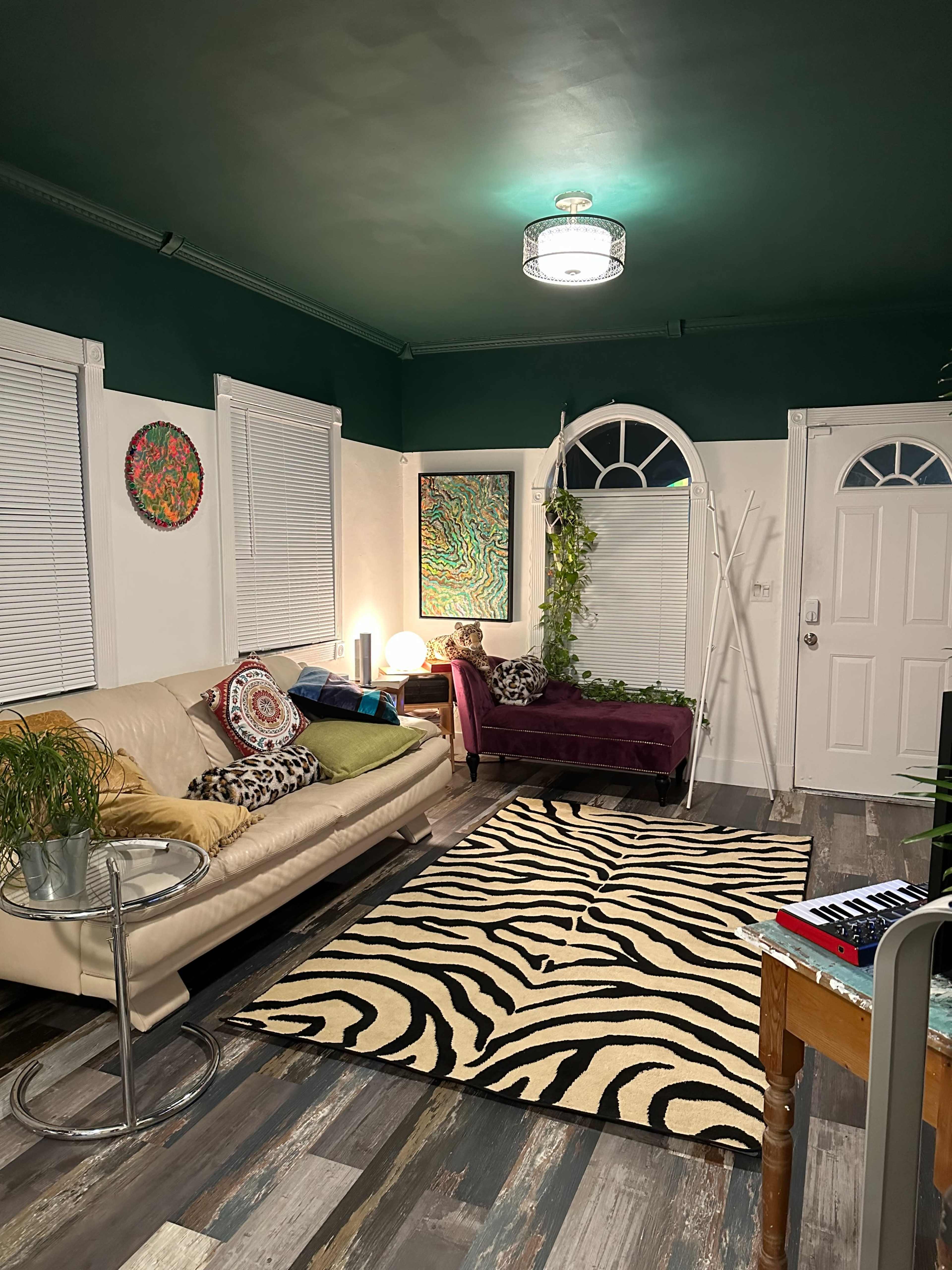 A cozy living room features a beige sofa, a purple lounge chair, a zebra-print rug, colorful wall art, and a door leading outside.
