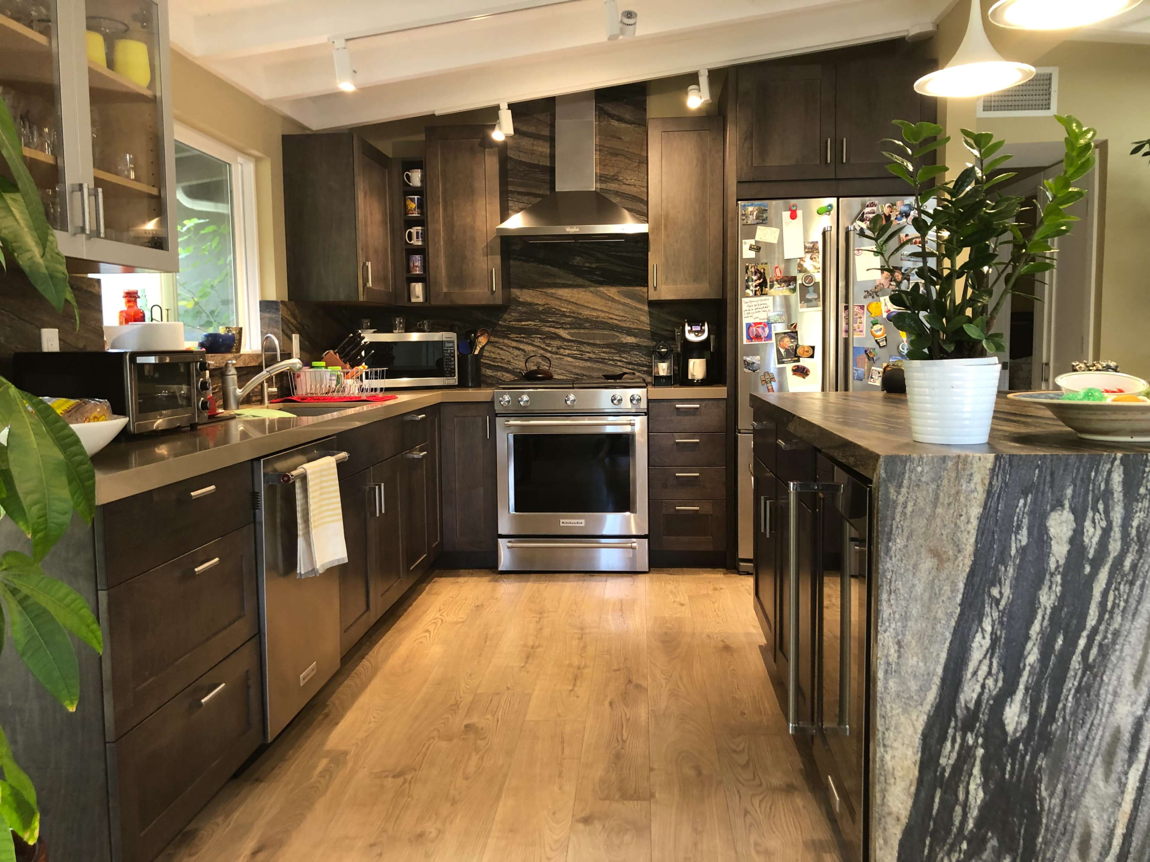 The image shows a modern kitchen with dark wooden cabinets, stainless steel appliances, and a stone island, complemented by indoor plants and a bright atmosphere.