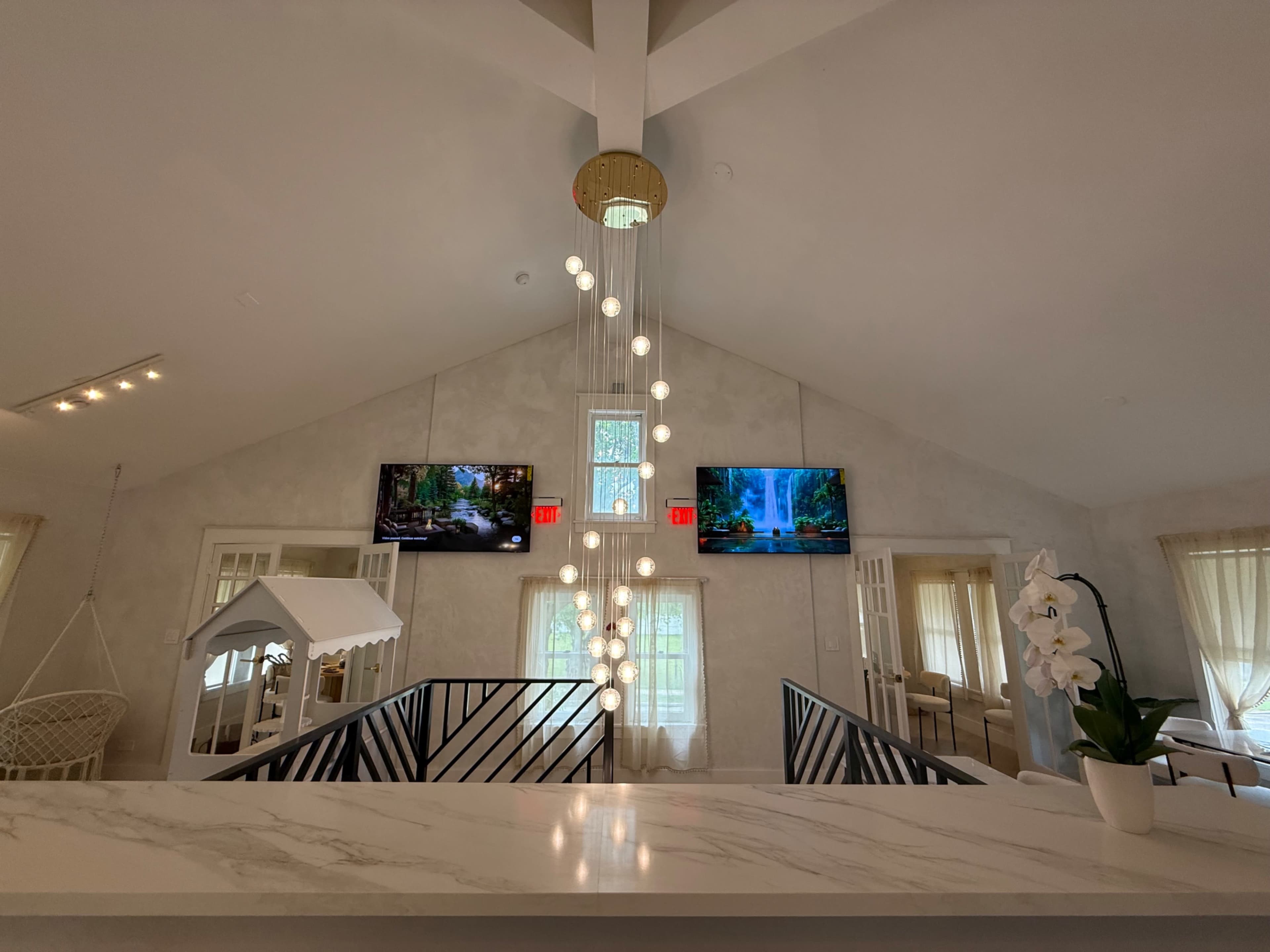The image shows a modern interior space with two wall-mounted televisions displaying nature scenes, an elegant chandelier, and a white countertop.